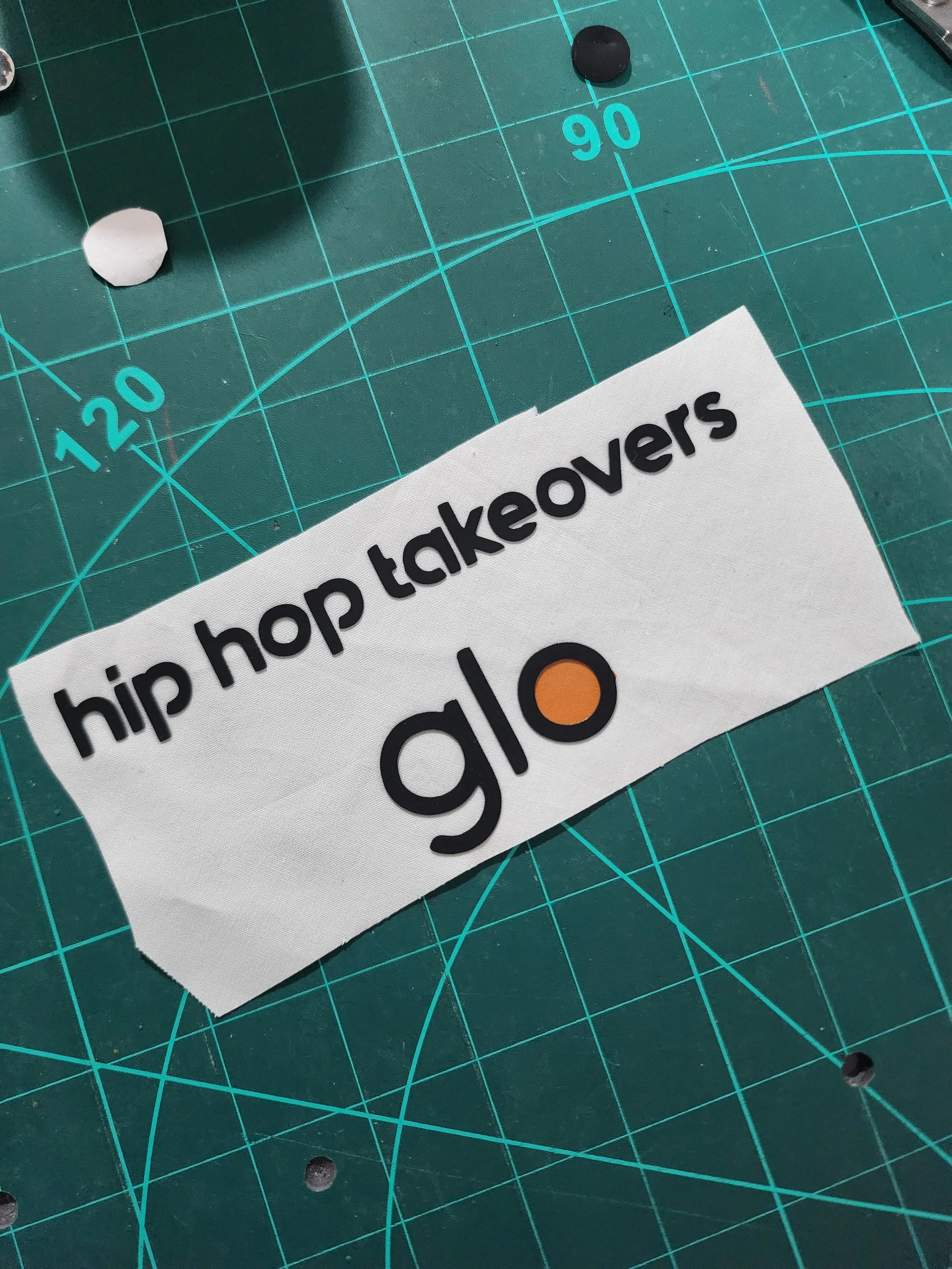 A fabric patch with the text 'hip hop takeovers' and a logo with the words 'glo' and a stylized orange circle on a green cutting mat with measurement grid lines.