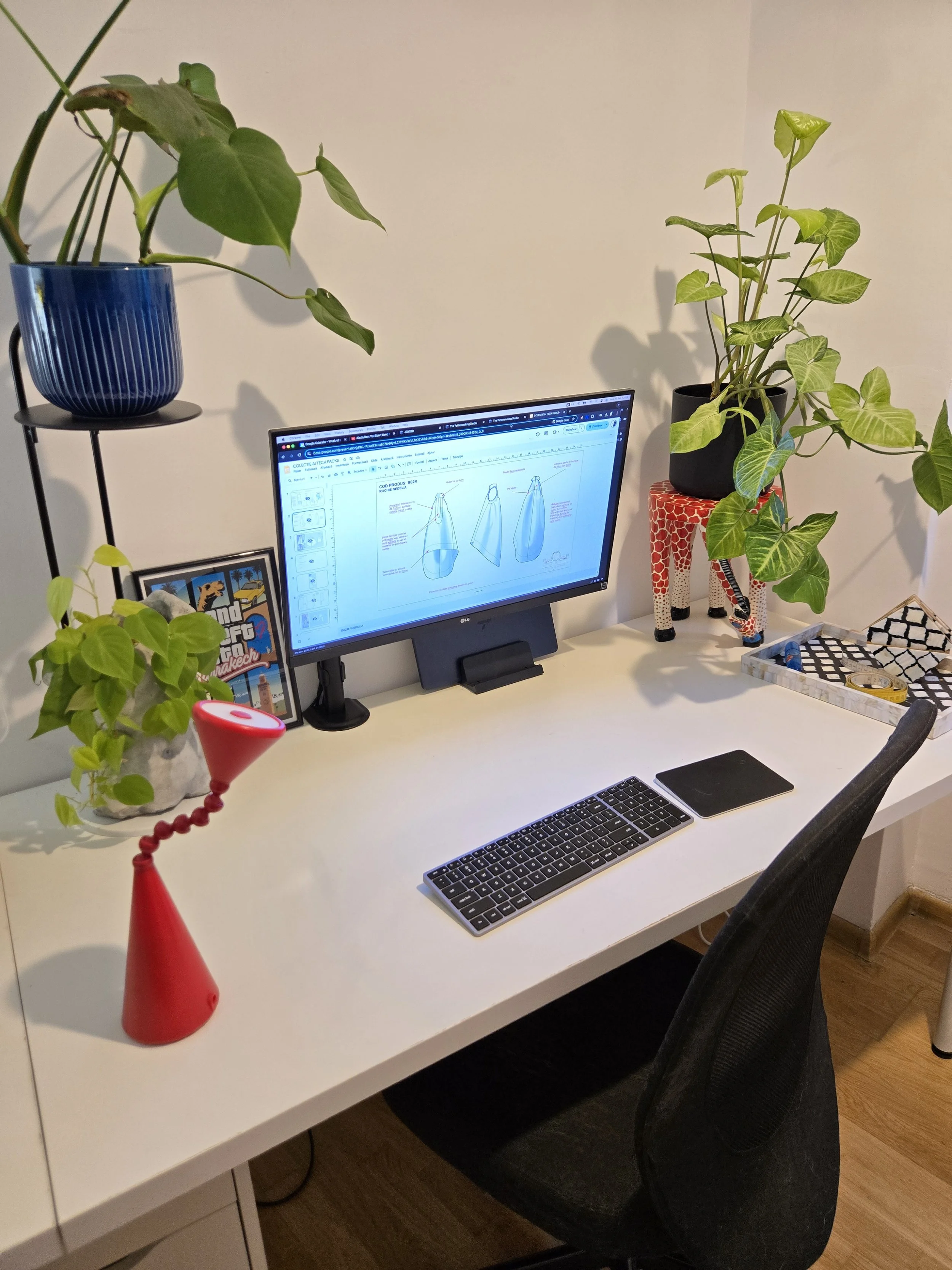 Home office desk setup with a white desk, black chair, computer monitor displaying design sketches, keyboard, and mouse. Decor includes several potted plants, a framed photo, a red decorative vase, and a jewelry tray, with natural light illuminating the workspace.