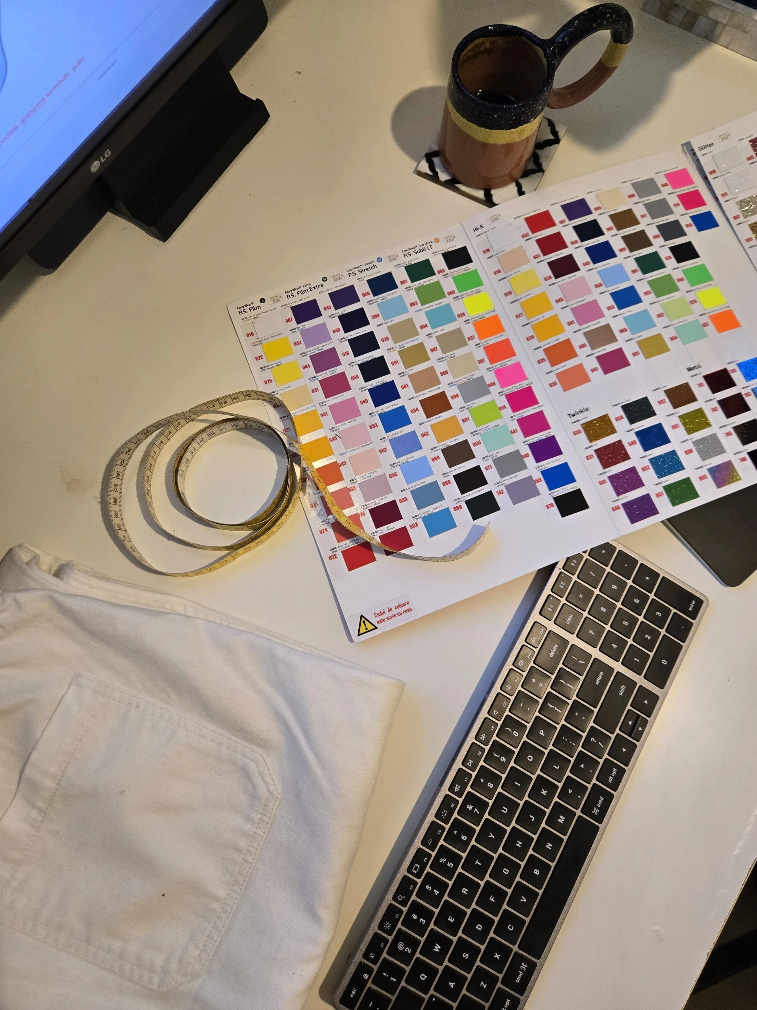 Color swatch chart, a flexible measuring tape, a white cloth, a coffee mug, a computer monitor, and a computer keyboard on a white desk.