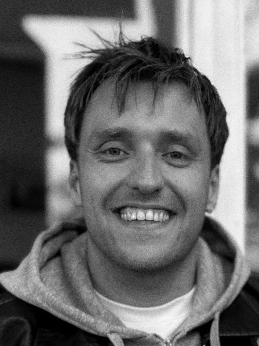 A black and white photo of a young man with short, tousled hair, smiling at the camera. He is wearing a hoodie and a leather jacket, standing indoors.