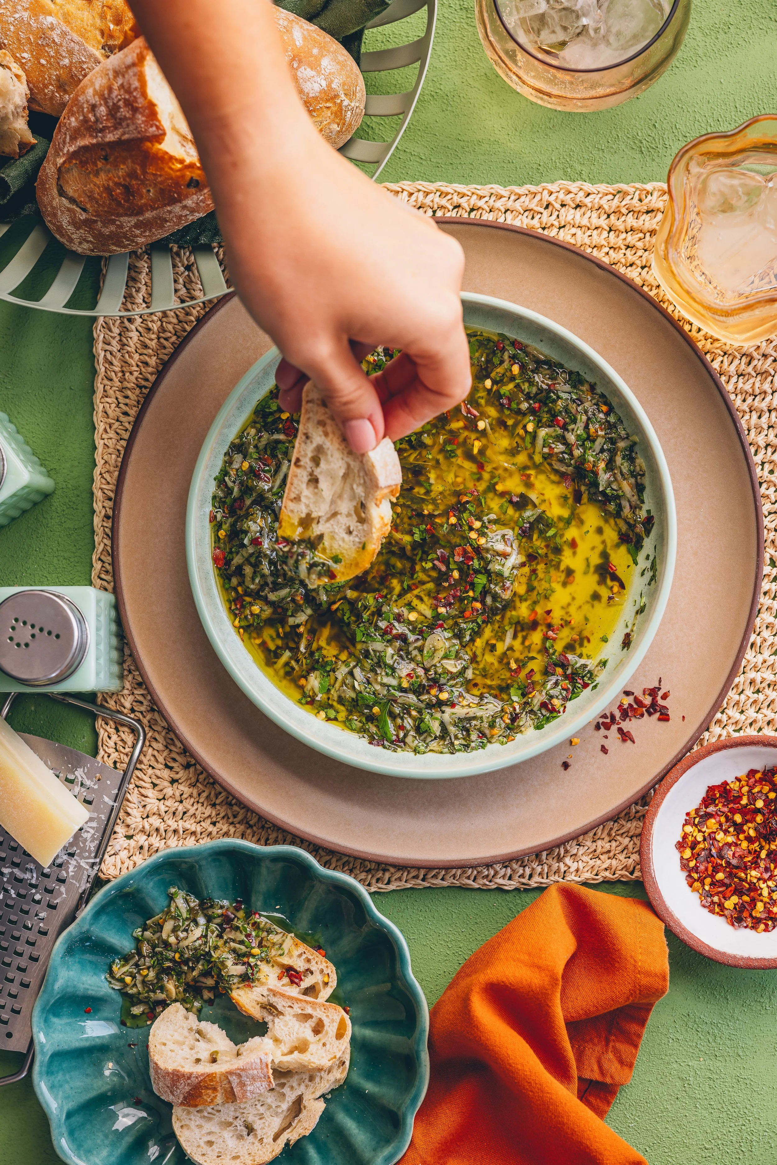 A person dipping a piece of bread into a bowl of chopped herbs, oil, and red pepper flakes on a green table. Surrounding the bowl are various bread pieces, glasses of iced drinks, and spice containers.
