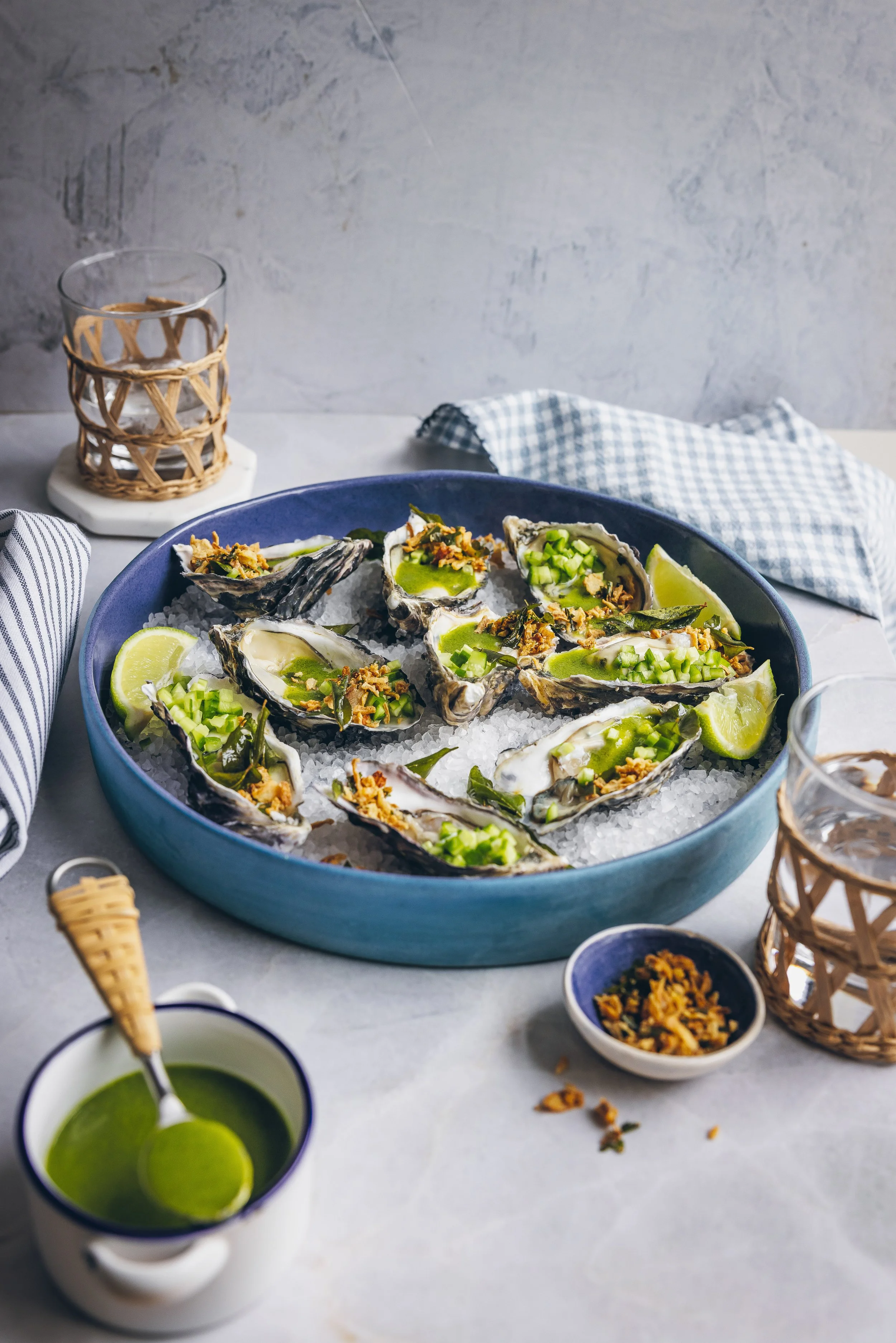 A blue bowl filled with fresh oysters topped with chopped green onions and herbs, served on ice with lime wedges, surrounded by small bowls of sauce, napkins, and glasses of drink on a white marble table.