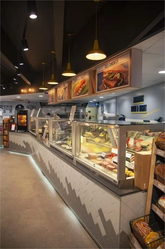 Inside a modern deli or sandwich shop with a glass display case filled with meats and cheeses, menu signs above advertising chicken salad and BLT sandwiches, and warm lighting fixtures hanging from the ceiling.