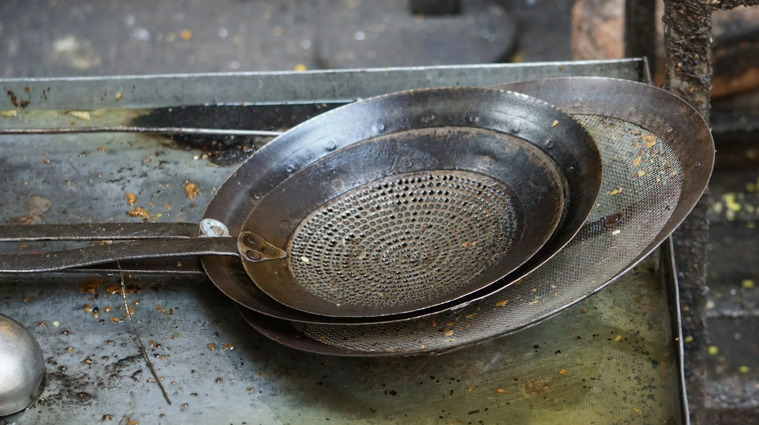 A rusty metal grill with small holes, resting on a dirty metal surface with scattered debris and food particles.