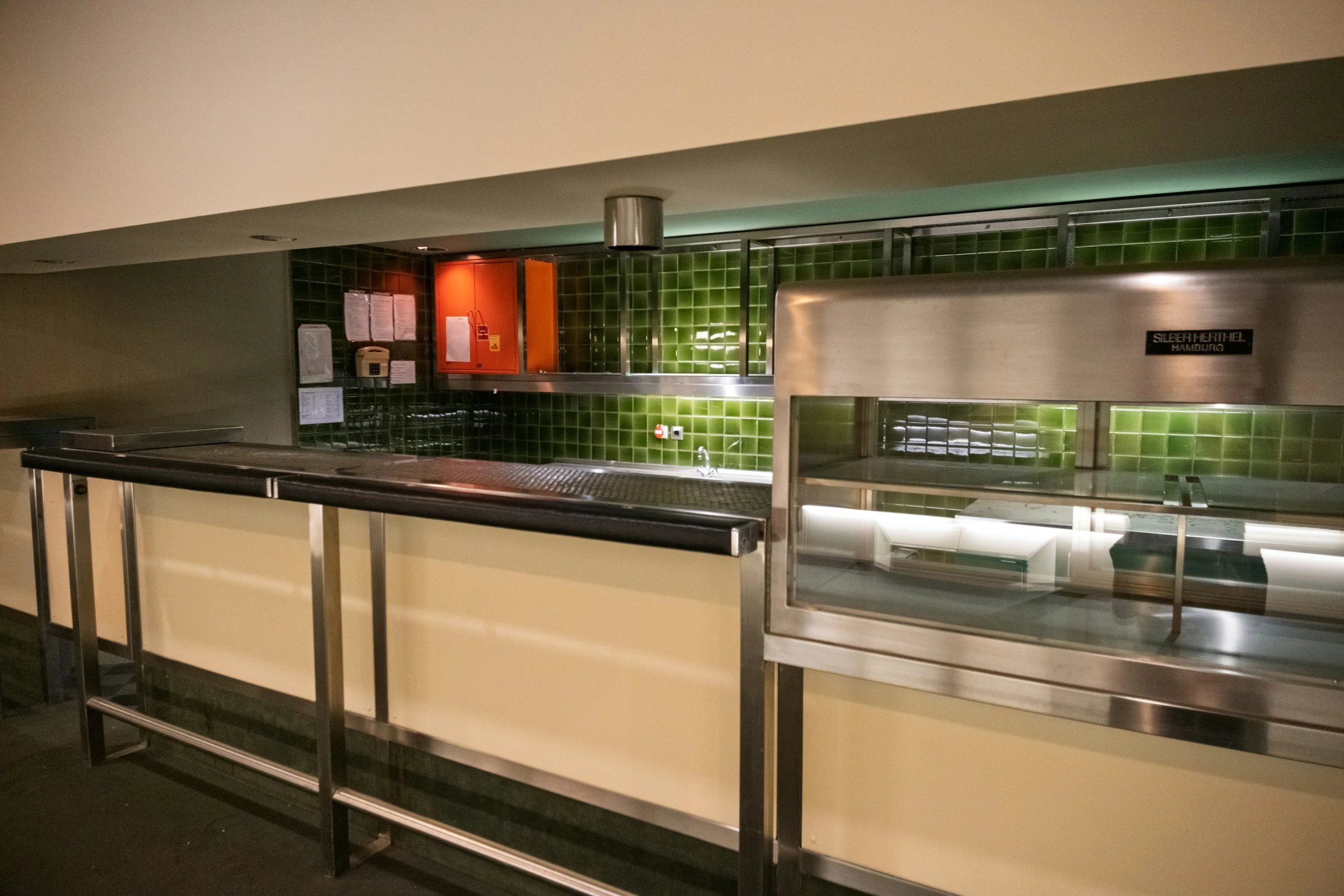 Empty cafeteria counter with green tiled wall in the background, orange and black fire safety equipment, electrical outlets, and stainless steel serving station labeled 'Silber Heribel Hamburg'.