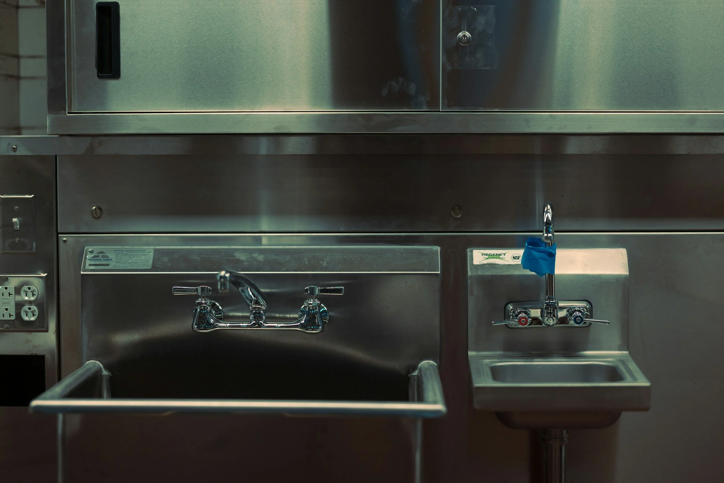 Stainless steel medical or laboratory sink with faucet, soap dispenser, and blue sanitizer bottle in a clinical setting.
