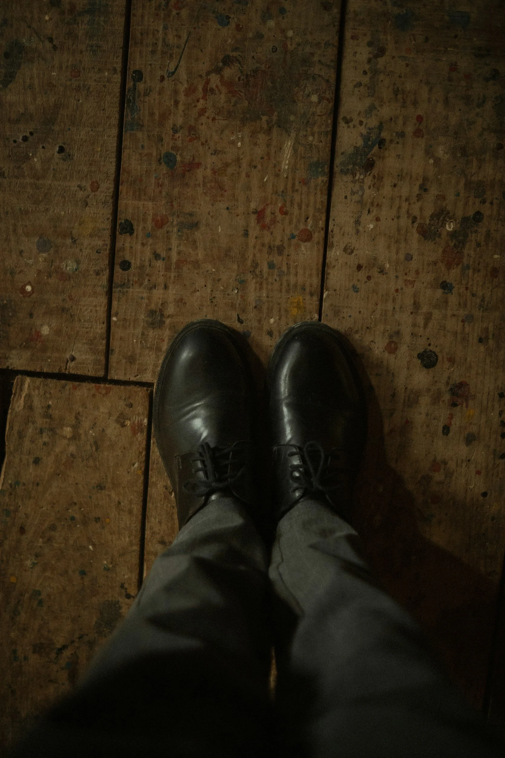 A person's feet wearing black shoes and gray pants standing on a worn wooden floor with paint splatters.