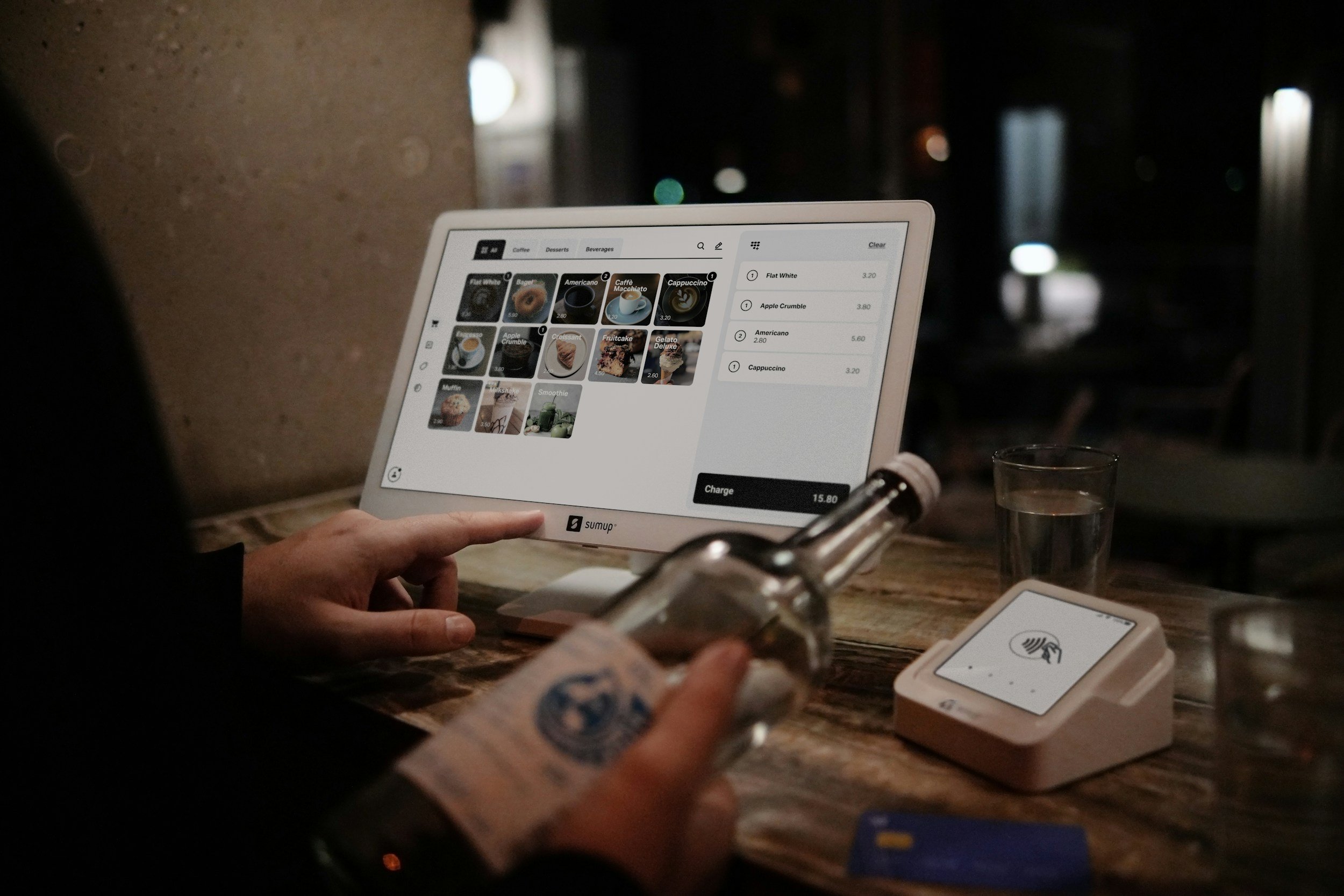 A person holding a tablet displaying a digital menu with various coffee options in a dimly lit cafe. There's a glass of water and a card reader on the table.