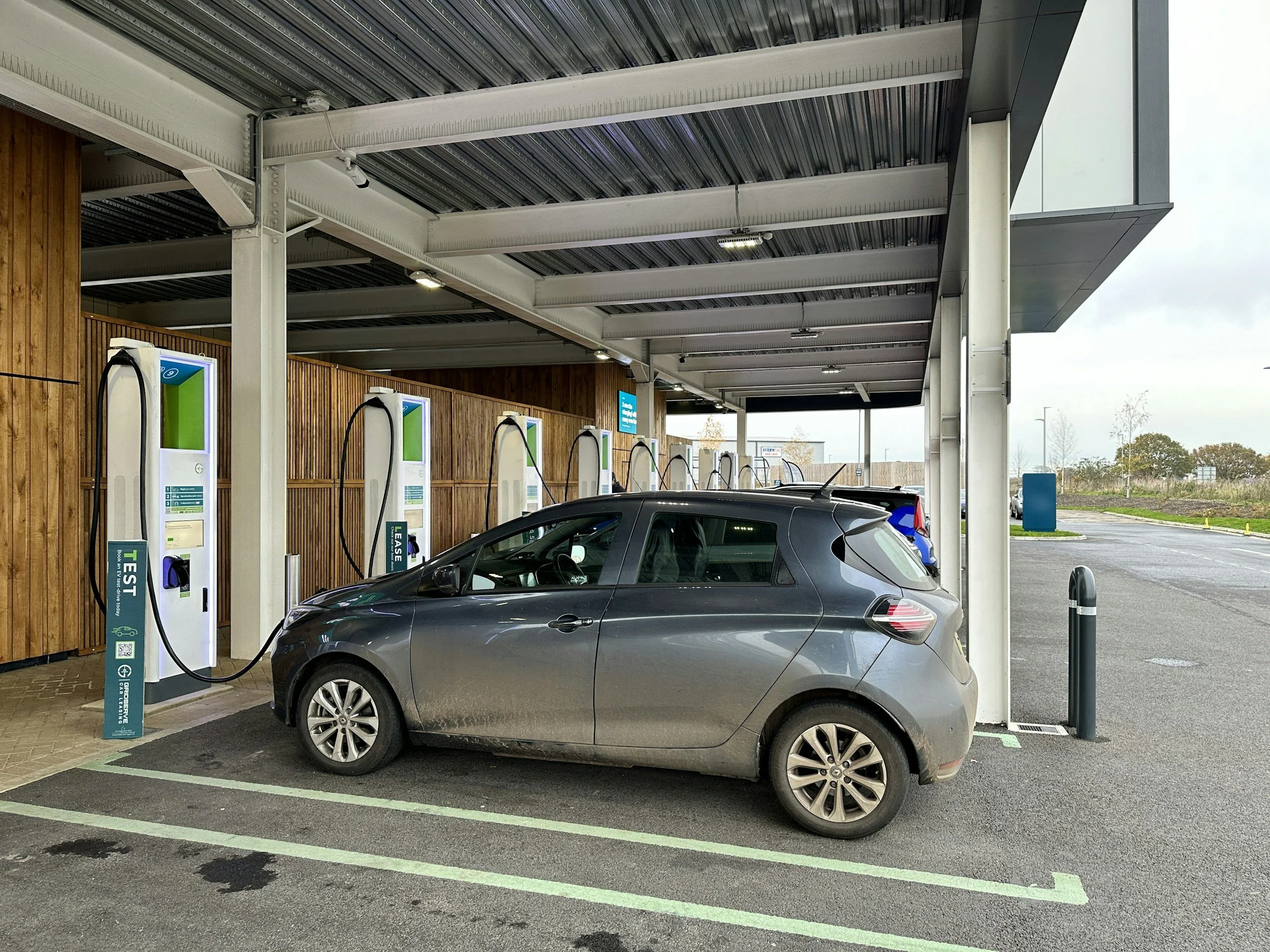 Electric vehicle charging station with a parked gray car, under a covered area with wooden and metal panels, outdoor parking lot, and cloudy sky.