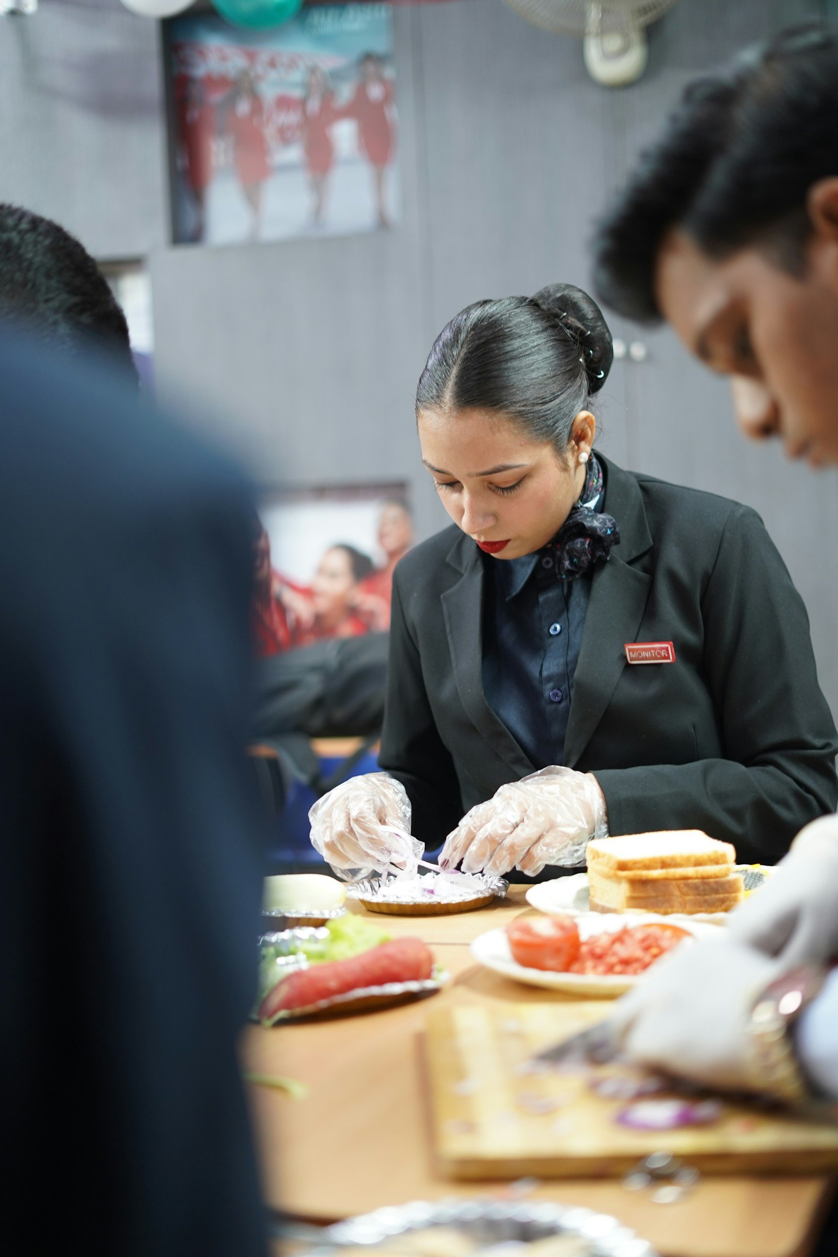 A woman dressed in a black suit with a name tag reading 'Monitor' is preparing food at a table, wearing plastic gloves. The table has various ingredients, including sliced tomatoes, bread, and vegetables. Several people are in the background, and there is a blurred poster on the wall.
