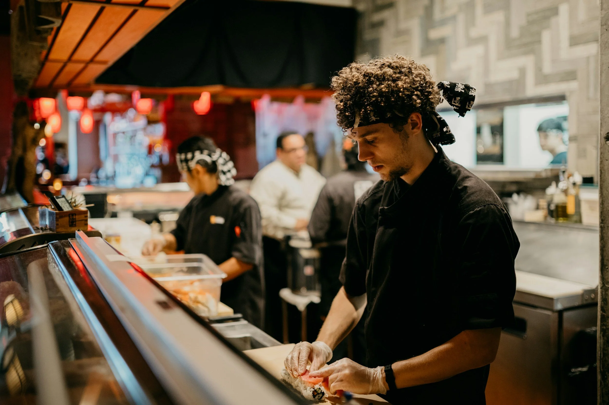 A sushi chef prepares sushi in a busy restaurant, with patrons and staff visible in the background.