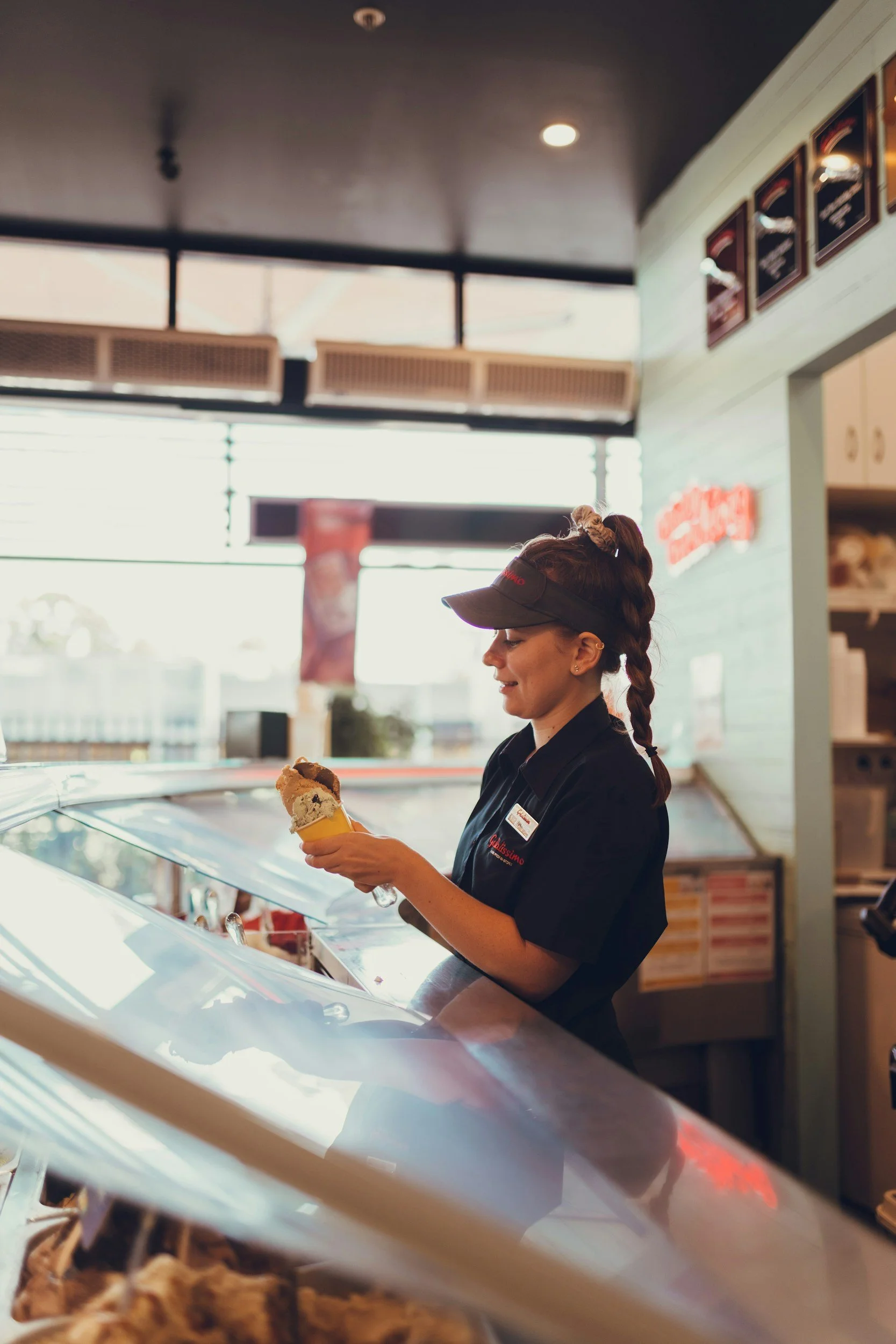 A woman with a dark uniform and a visor cap serving ice cream in a cone inside an ice cream shop.