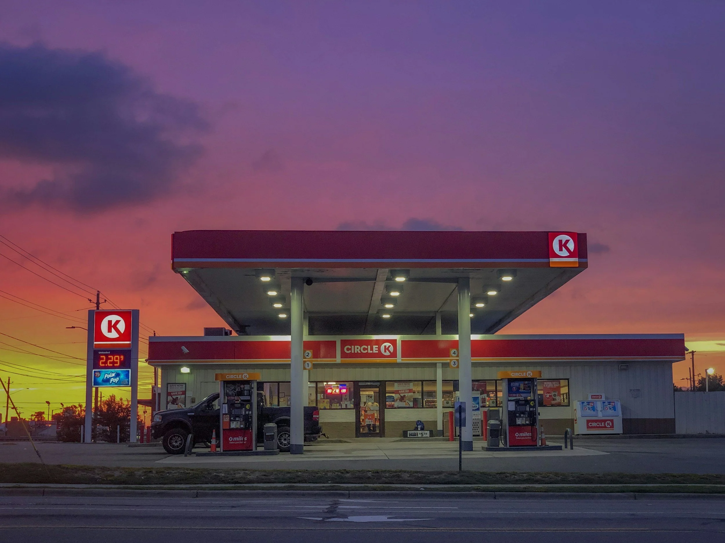 A Circle K convenience store and gas station at sunset with a colorful sky, multiple gas pumps, a sign showing the price of unleaded gasoline at $2.29, and a few parked cars.