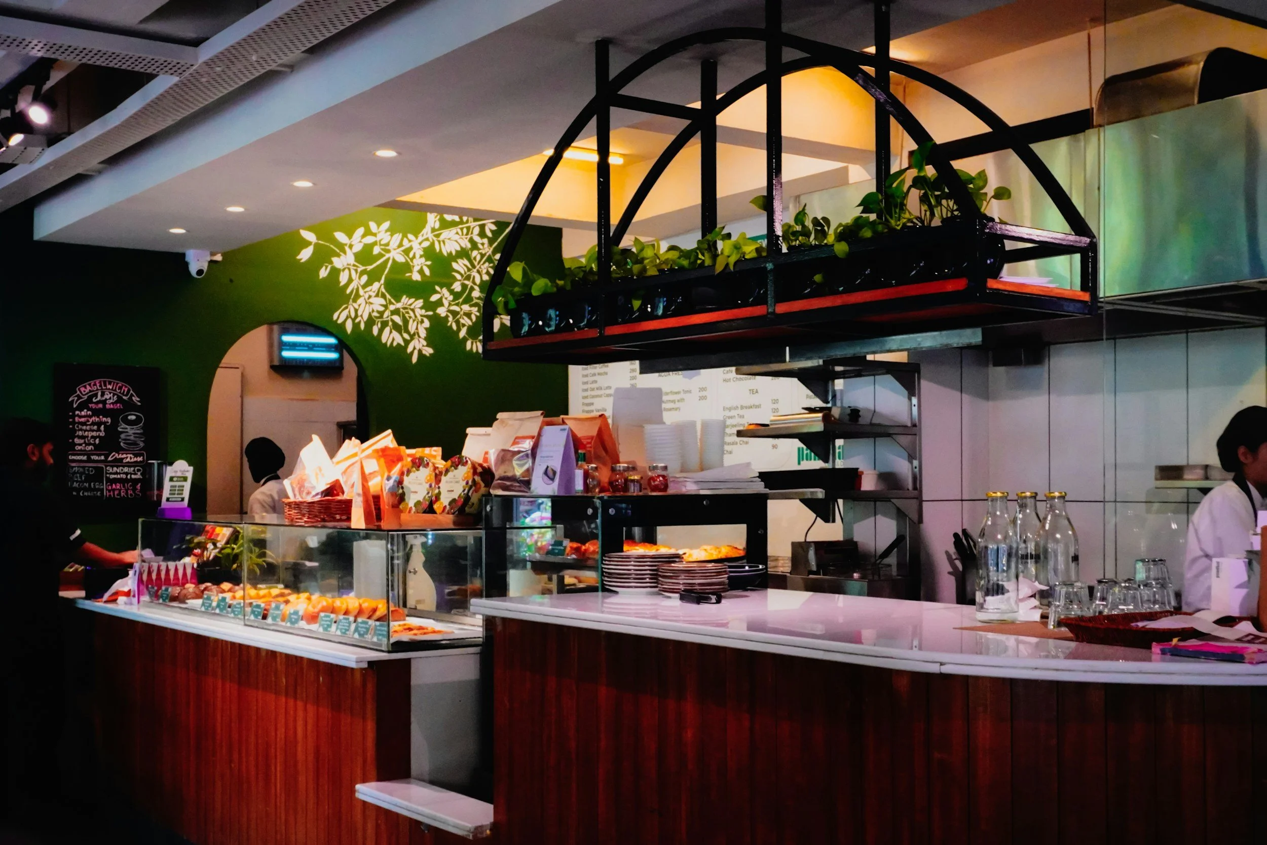 Inside a cafe with a counter displaying desserts, a green wall with a white leafy pattern, and a black metal hanging shelf with plants.