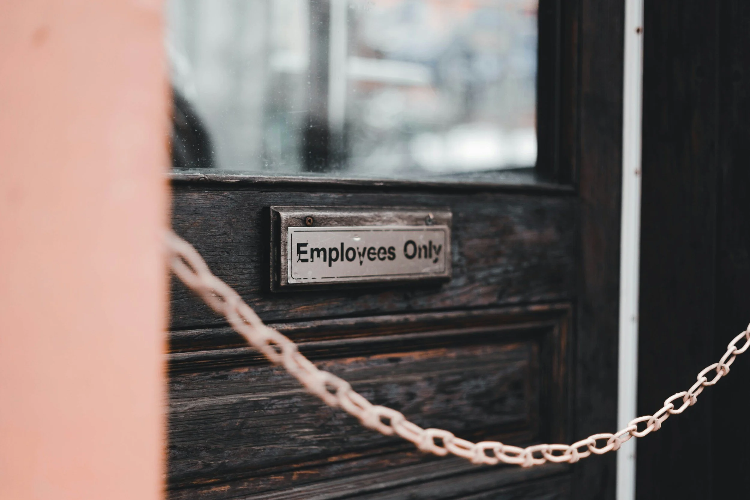 Close-up of a wooden door with a metal sign that reads 'Employees Only' and a pink chain barrier in front of it.