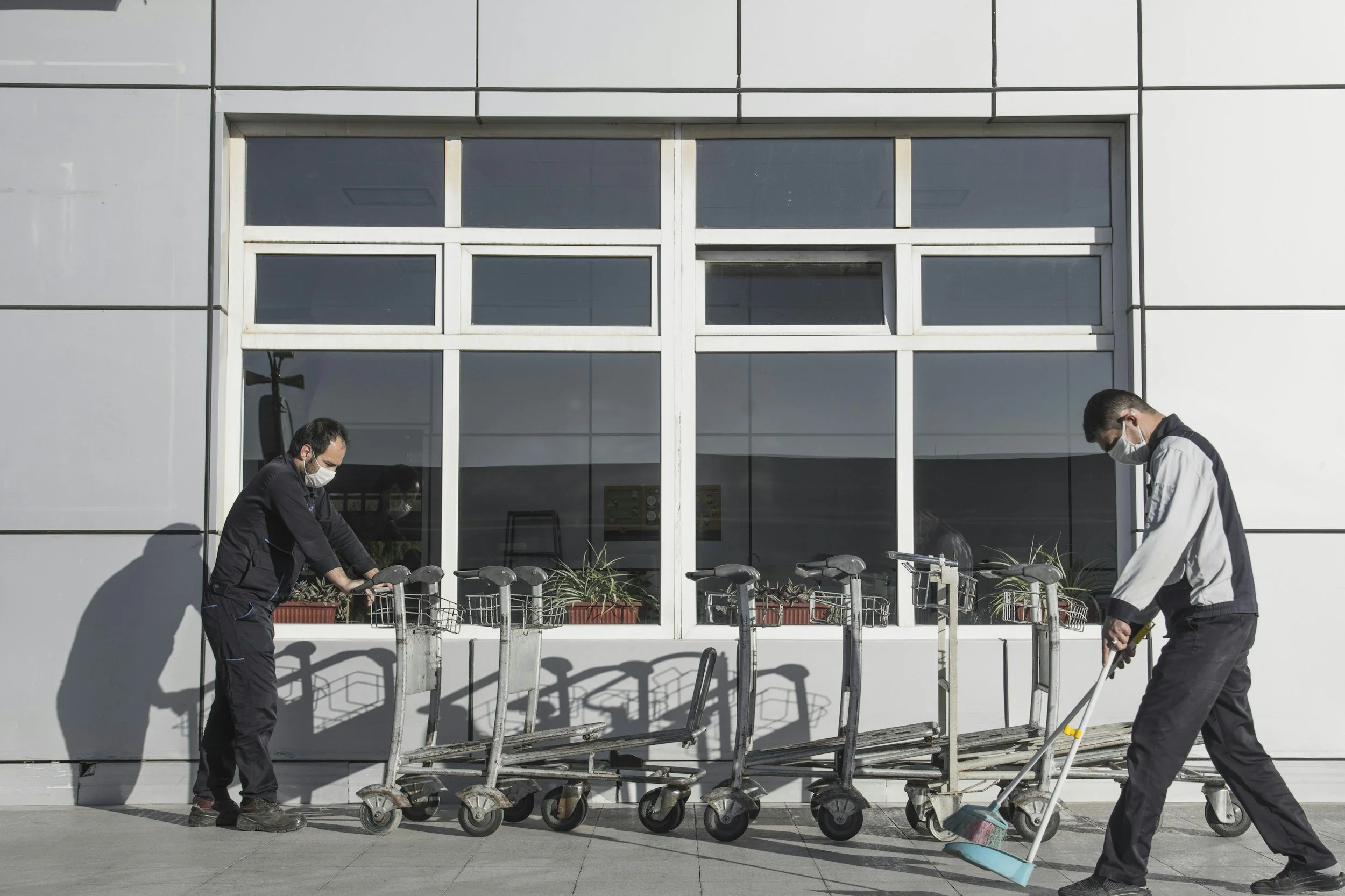 Two men wearing face masks cleaning shopping carts outside a modern building with large windows and white paneling.