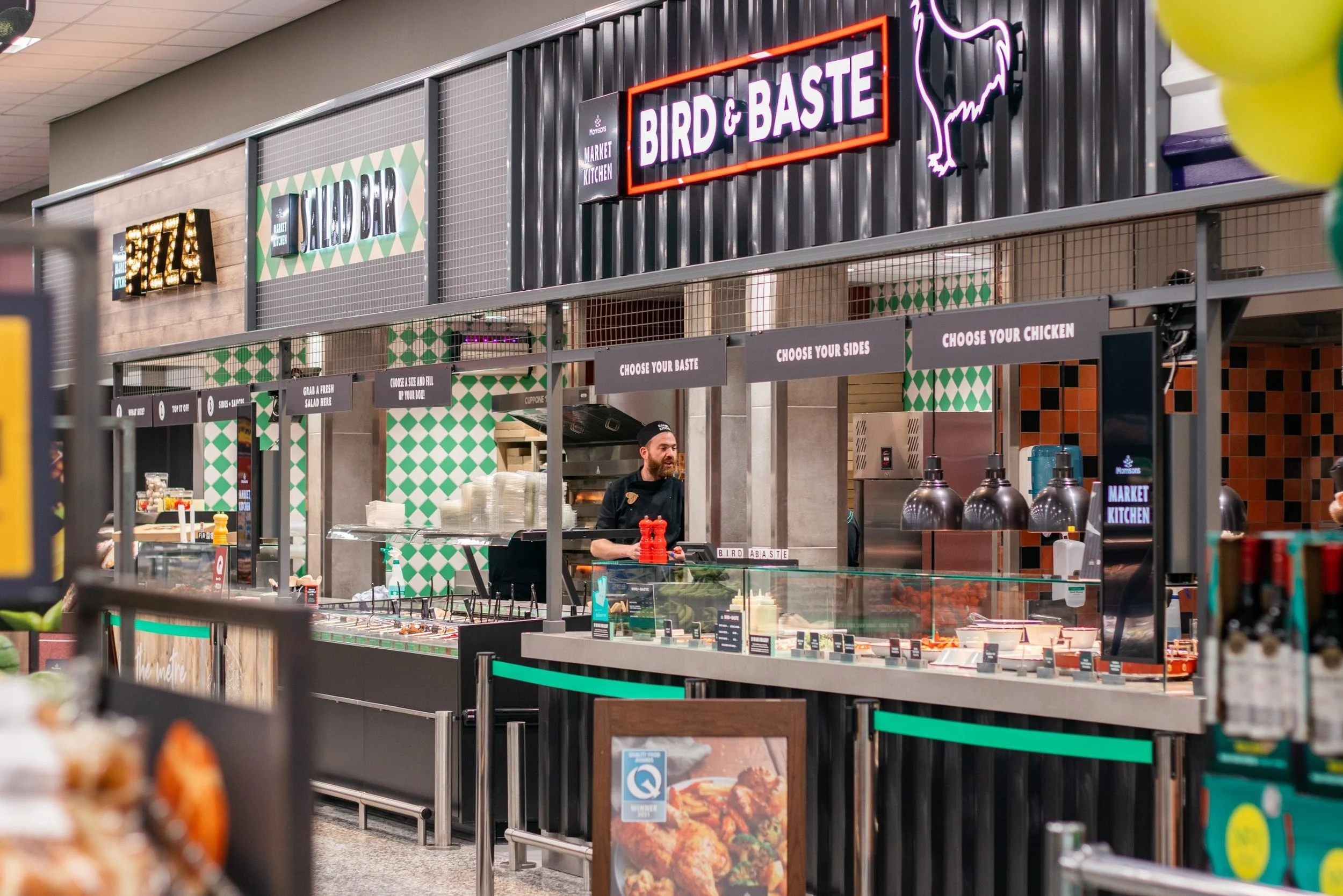 View of a food counter at a supermarket with signs indicating options for chicken, salad, sides, and so on. The sign reads 'Bird & Baste' with neon lighting and a chicken illustration. Another sign reads 'Sl-a-dor' in marquee lights. A man behind the counter is preparing food, and there are various food items and condiment bottles on display.