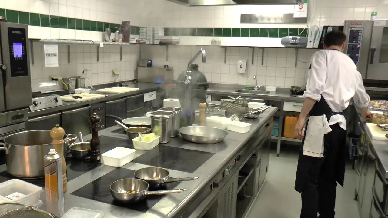 Commercial kitchen with various cooking utensils, pots, pans, and ingredients on stainless steel countertops. A chef in a white shirt and black apron is preparing food at a station.