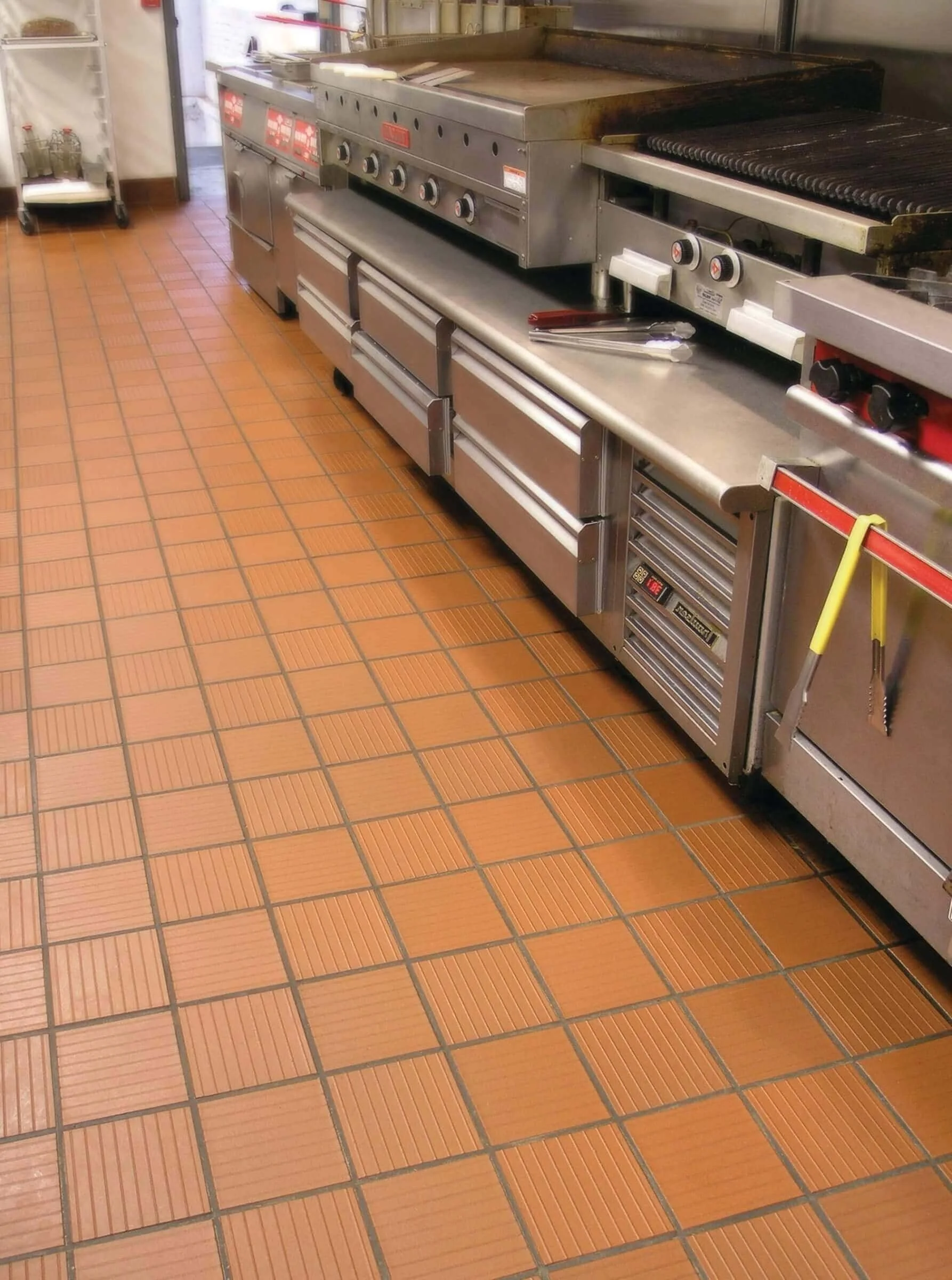 Commercial kitchen with a stainless steel stove, oven, and grill, situated on a tiled floor. There are utensils piled on the counter and a cart with glass bottles nearby.