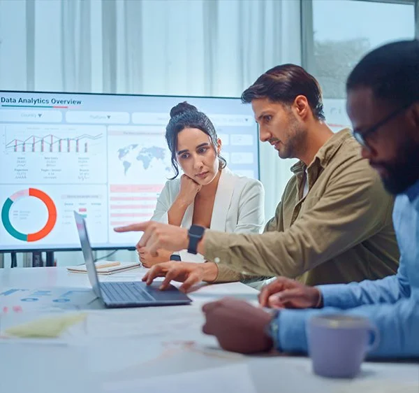 Three professionals working together in a meeting room with a large screen displaying data analytics charts and graphs.