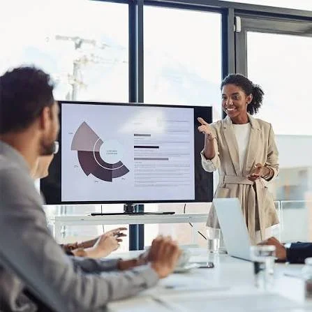 A woman presenting a business report on a large screen to a group of colleagues in a modern office conference room.