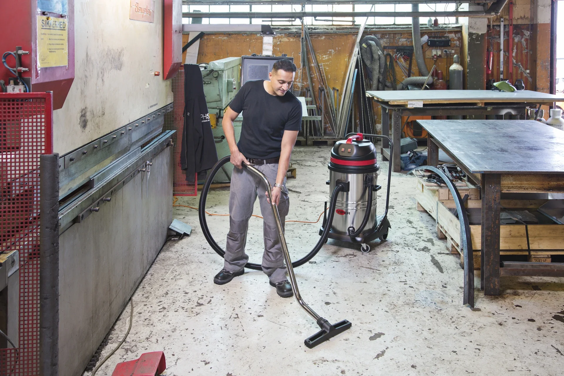 A man vacuuming a workshop or industrial space with machinery and workbenches in the background.