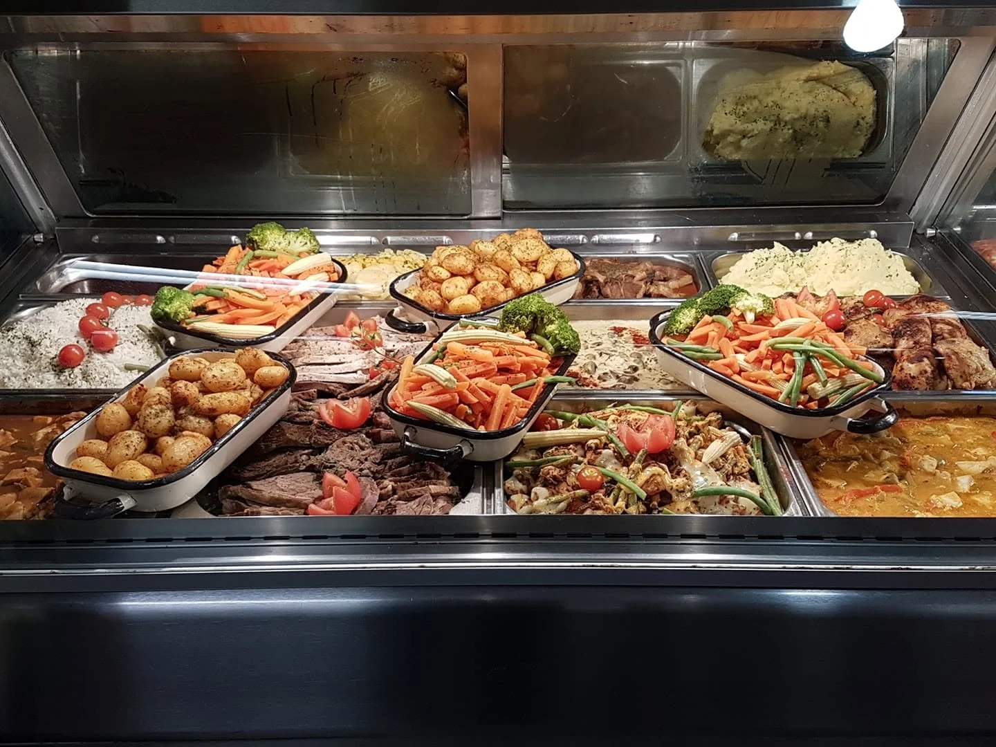 Variety of cooked vegetables, potatoes, meat, and rice dishes in a buffet display with metal trays and glass sneeze guard.
