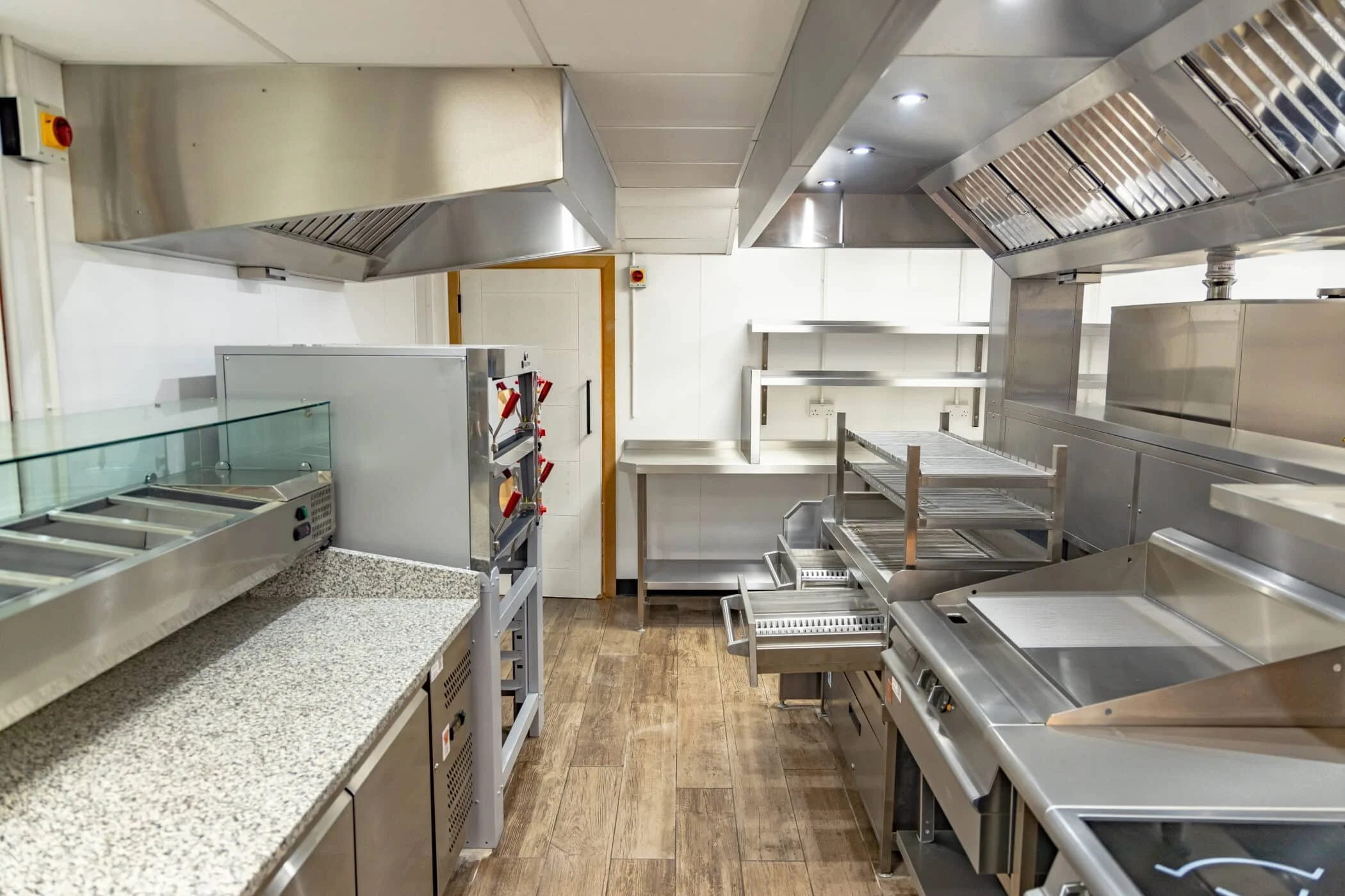 Commercial kitchen with stainless steel appliances and shelves, granite countertop, wooden floor, and white tiled walls.