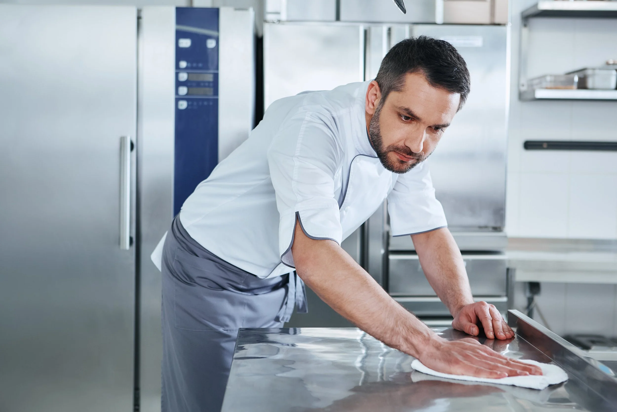 Man in a white chef coat preparing meat on a stainless steel counter in a commercial kitchen.
