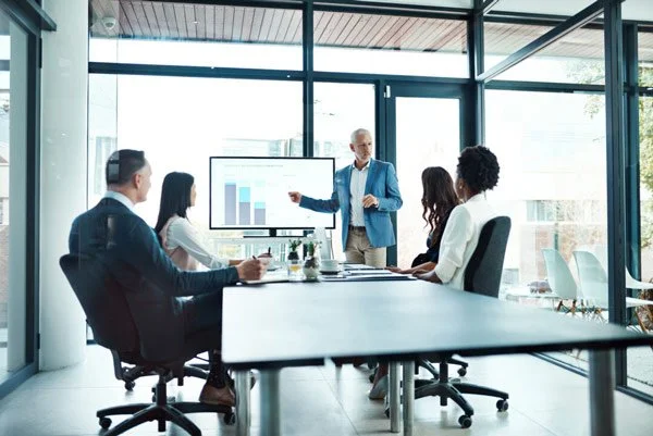 Business meeting with five people in a modern office conference room, one person presenting data on a large screen.