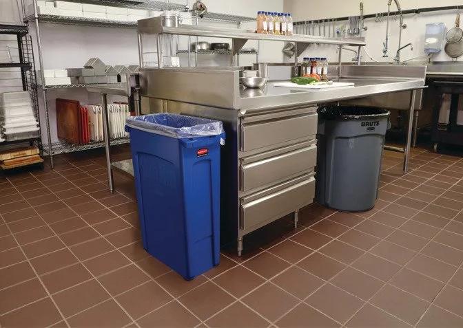 Commercial kitchen with stainless steel countertops and equipment, blue recycling bin, and gray trash can on brown tiled floor.
