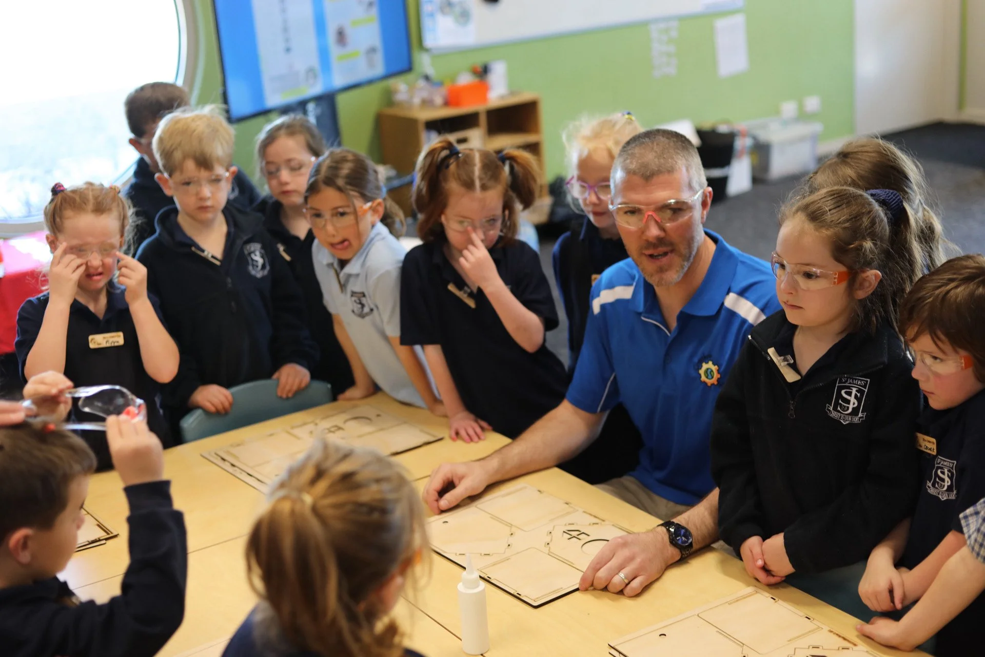 A group of elementary school children gathered around a teacher, who is demonstrating a science experiment or activity using wooden boards and a bottle of glue on a classroom table.