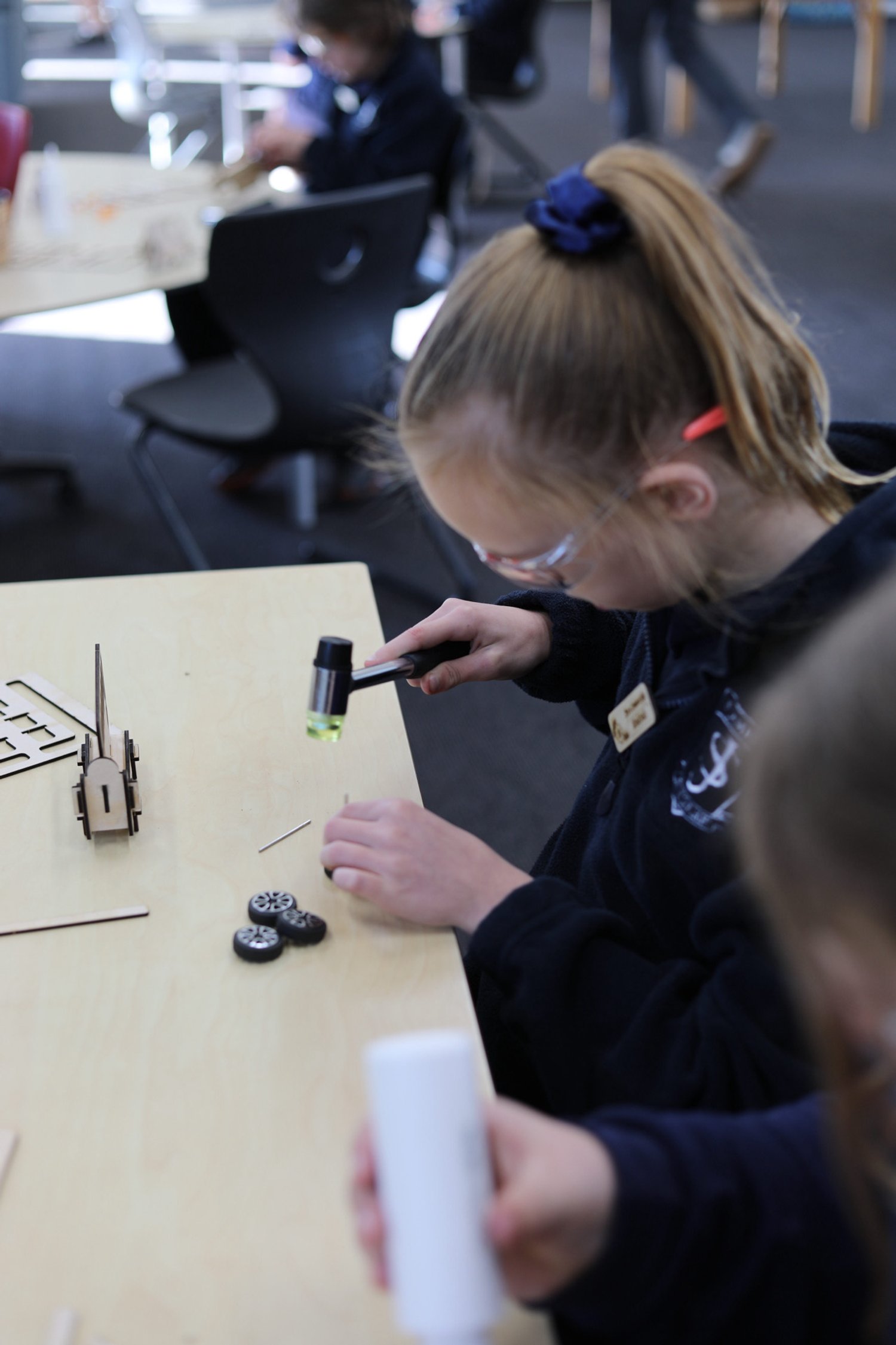 A young girl with glasses and a ponytail using a flashlight to examine small black toy wheels on a wooden table in a classroom.