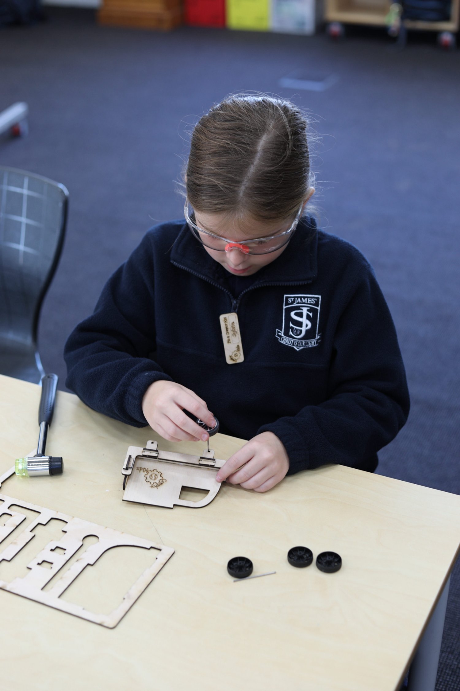 A young girl in safety glasses and a dark school uniform is working on assembling a small wooden model at a table. The table has additional wooden parts, a screwdriver, and three black wheels.