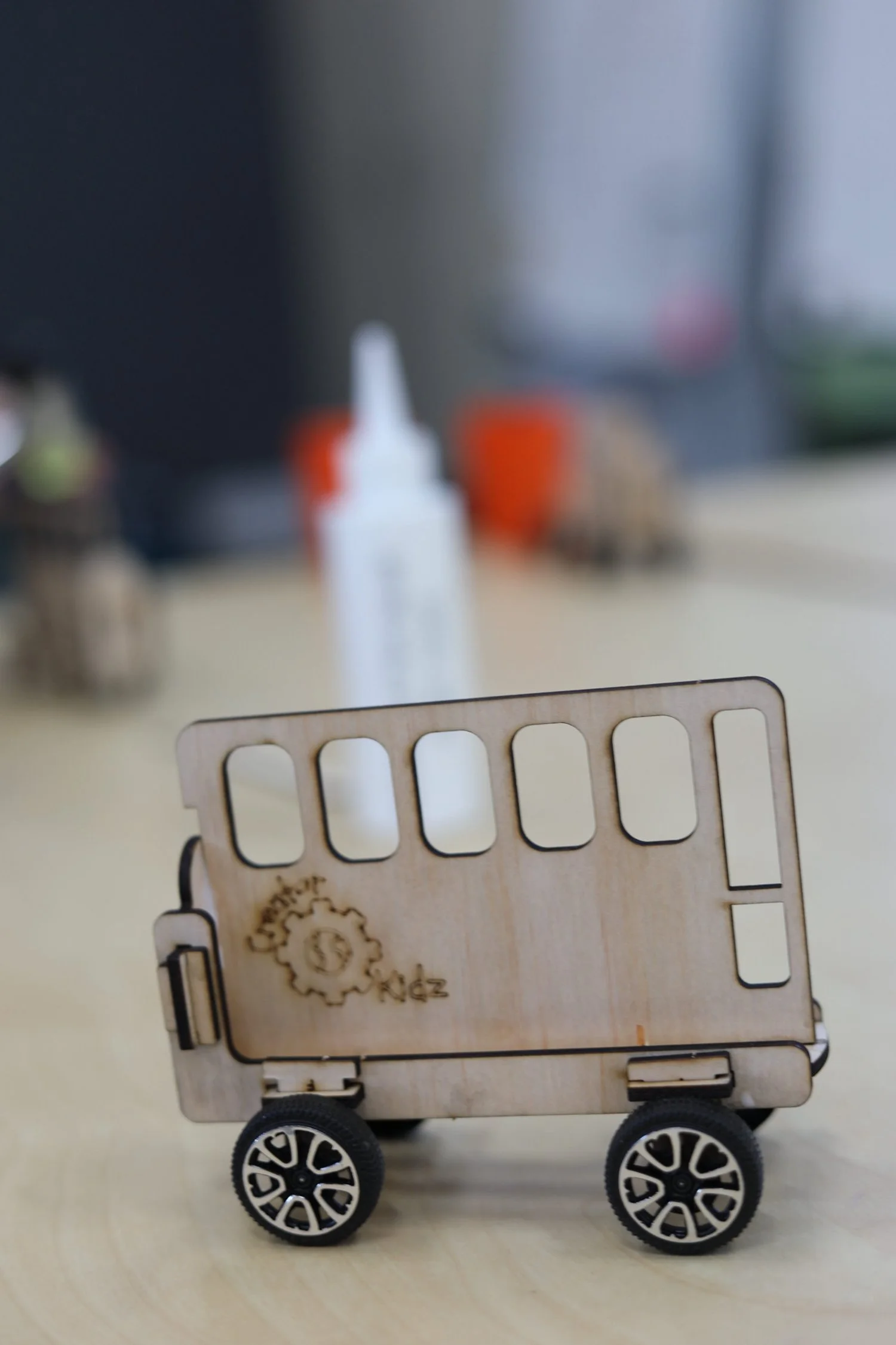 A small wooden toy bus with four black and white wheels, featuring a cutout window design, sitting on a wooden surface with a blurred background including a white squeeze bottle and other objects.