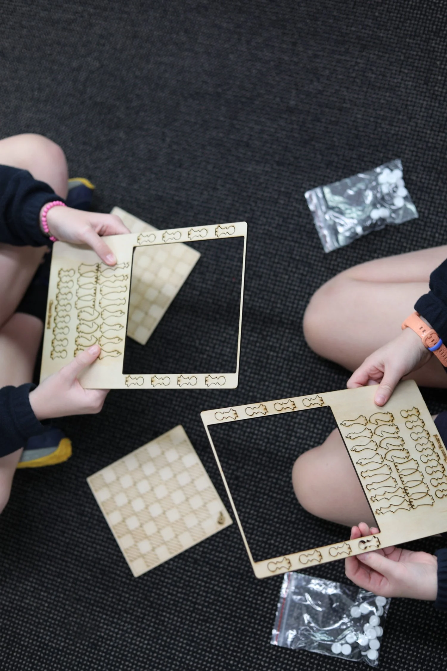 Two children sitting on a dark carpeted floor, each holding a square wooden frame with fish-shaped cutouts along the edges, above a chessboard and a small bag of checkers.