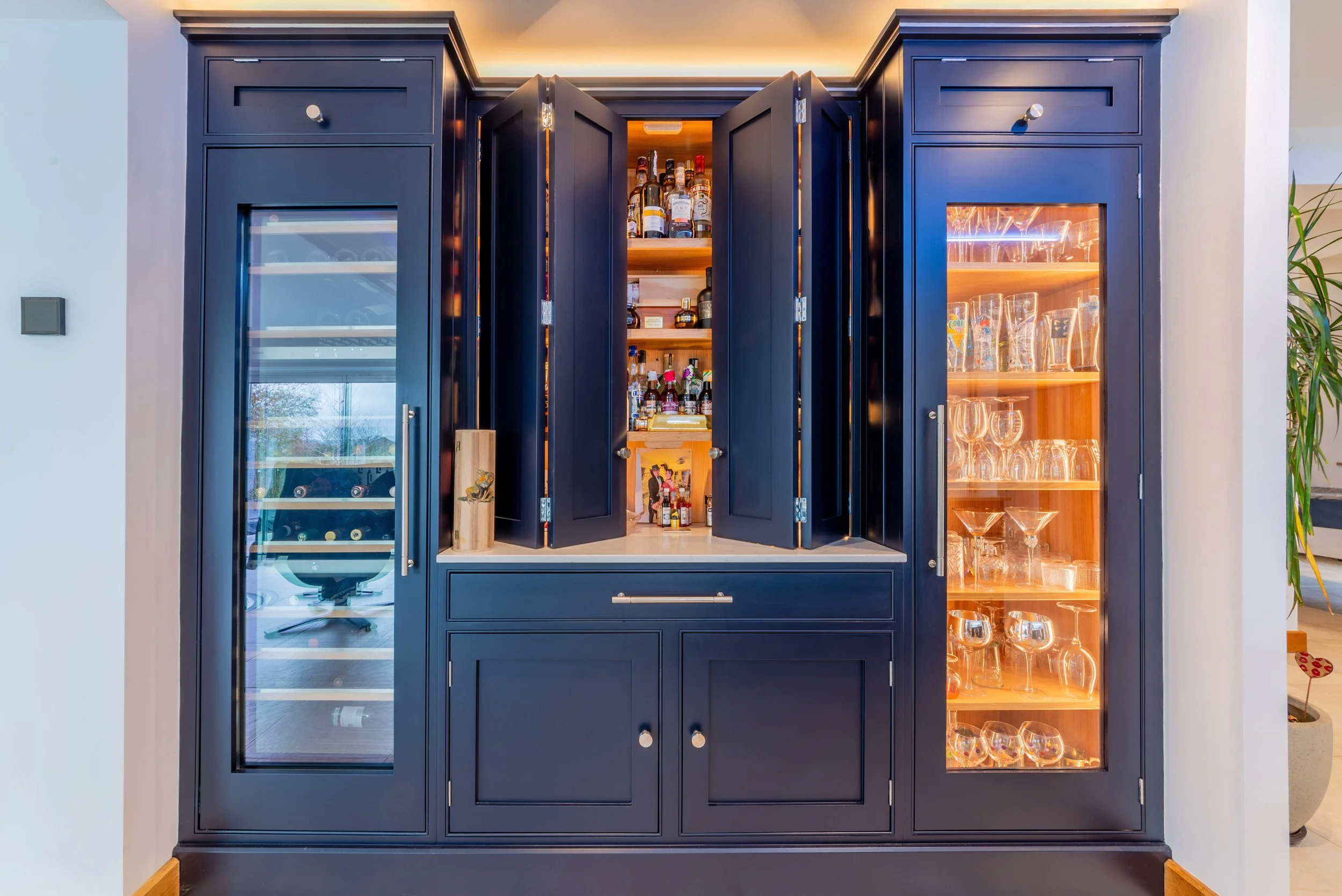 A built-in navy blue bar cabinet with glass doors on each side and open central doors showing bottles of alcohol, mixers, and glassware inside, in a modern interior.