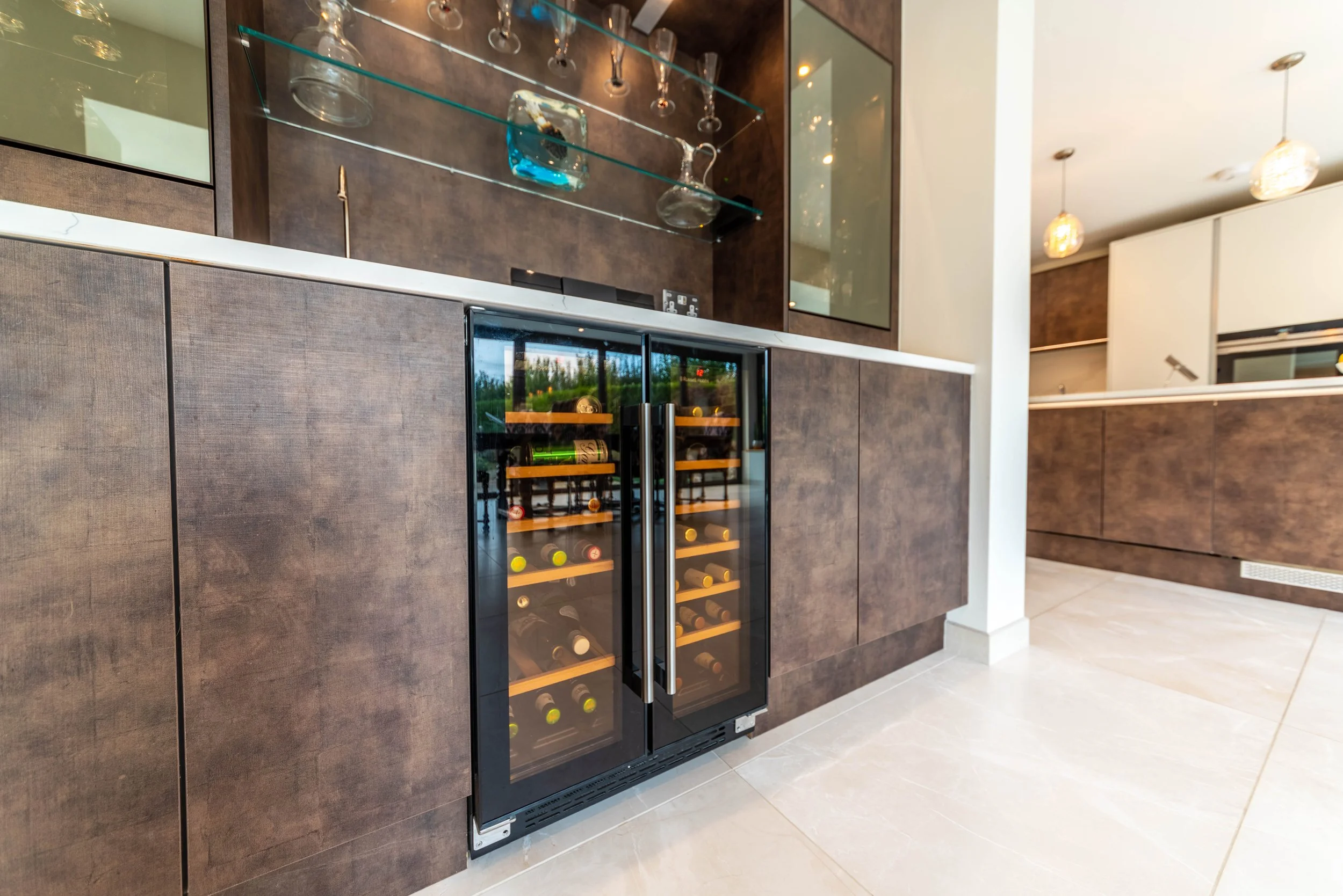 Drinkware displayed on glass shelves above a wine refrigerator in a modern kitchen with brown cabinetry and beige tiled floor.