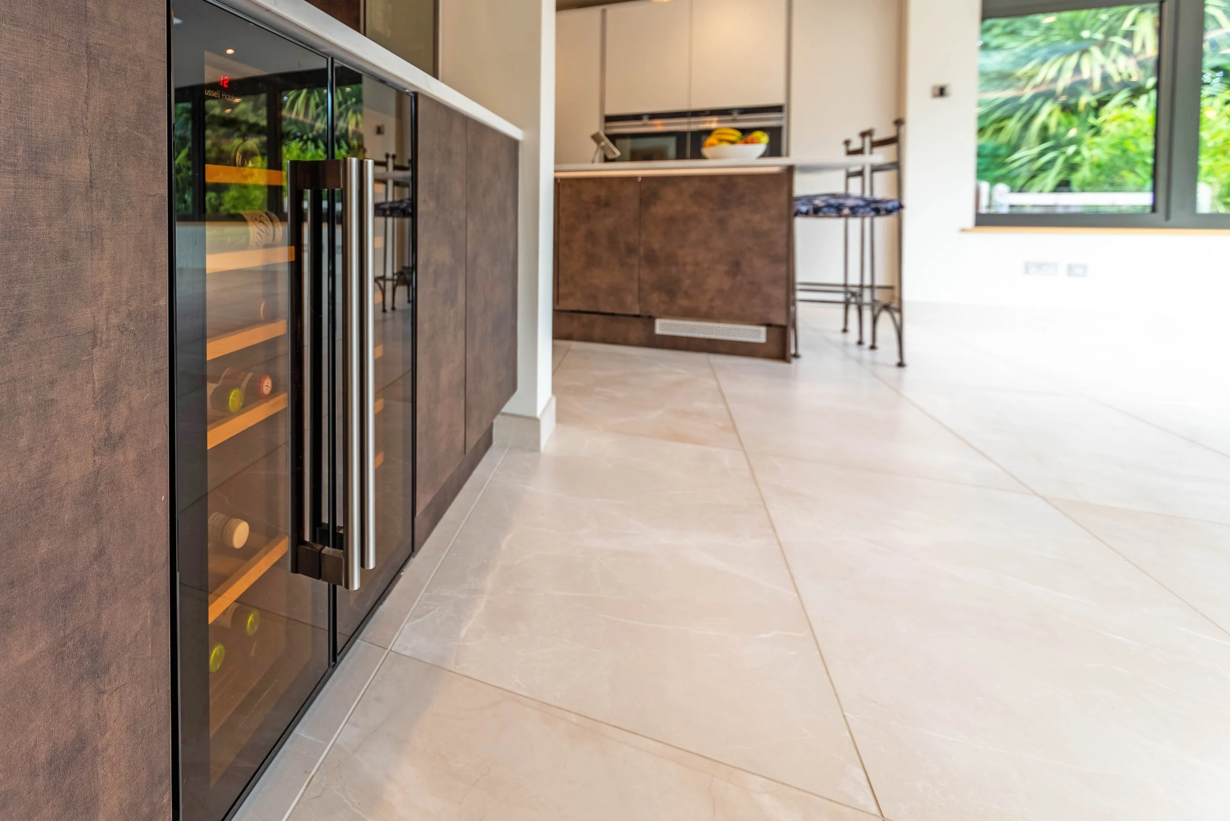 Modern kitchen with a wine cooler, beige tiled floor, brown cabinets, a kitchen island, and a window showing green outdoor foliage.
