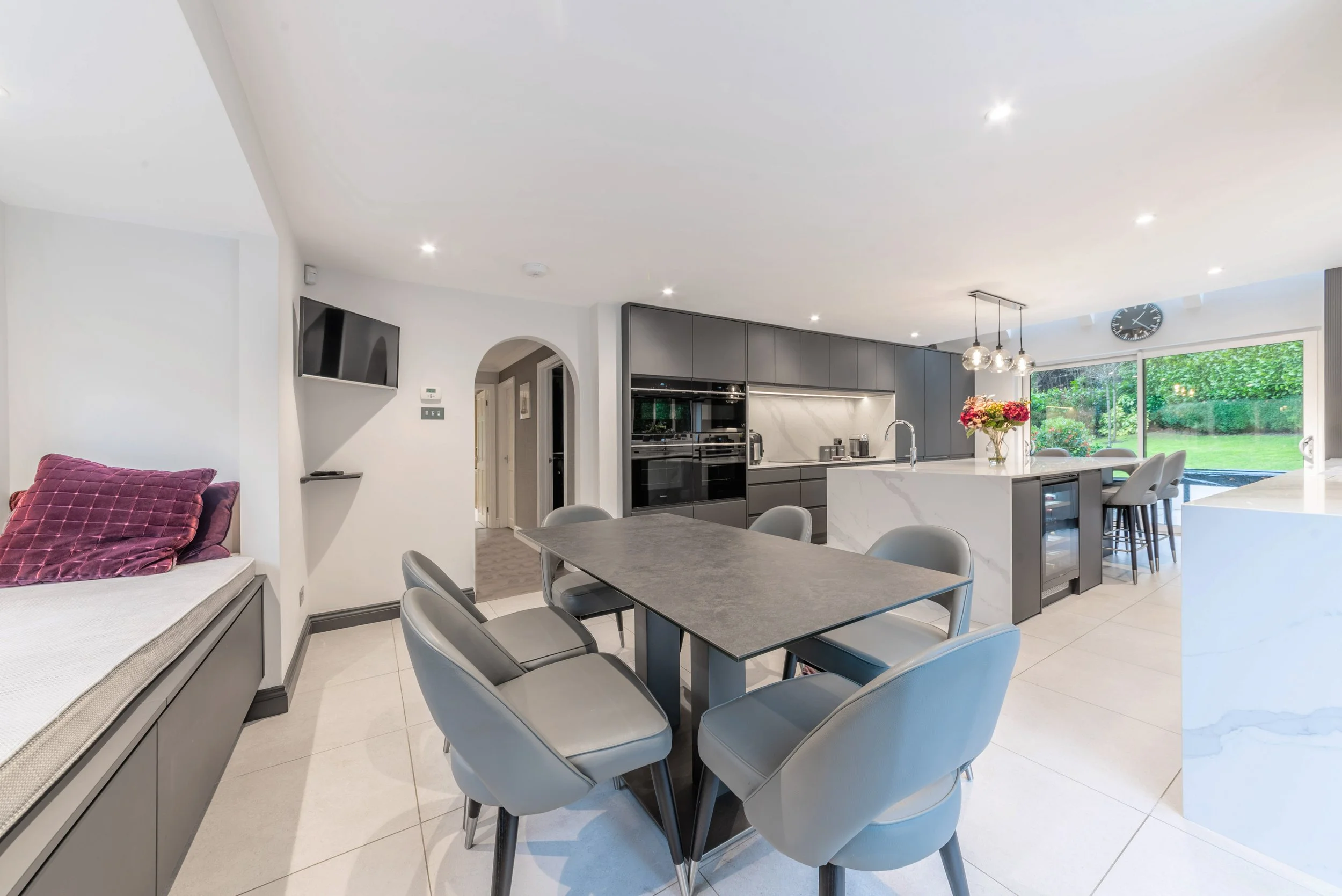 Modern open-plan kitchen and dining area with gray cabinets, a black dining table with six gray chairs, and a kitchen island with flowers, a walk-in closet with outdoor view, and a wall-mounted TV.