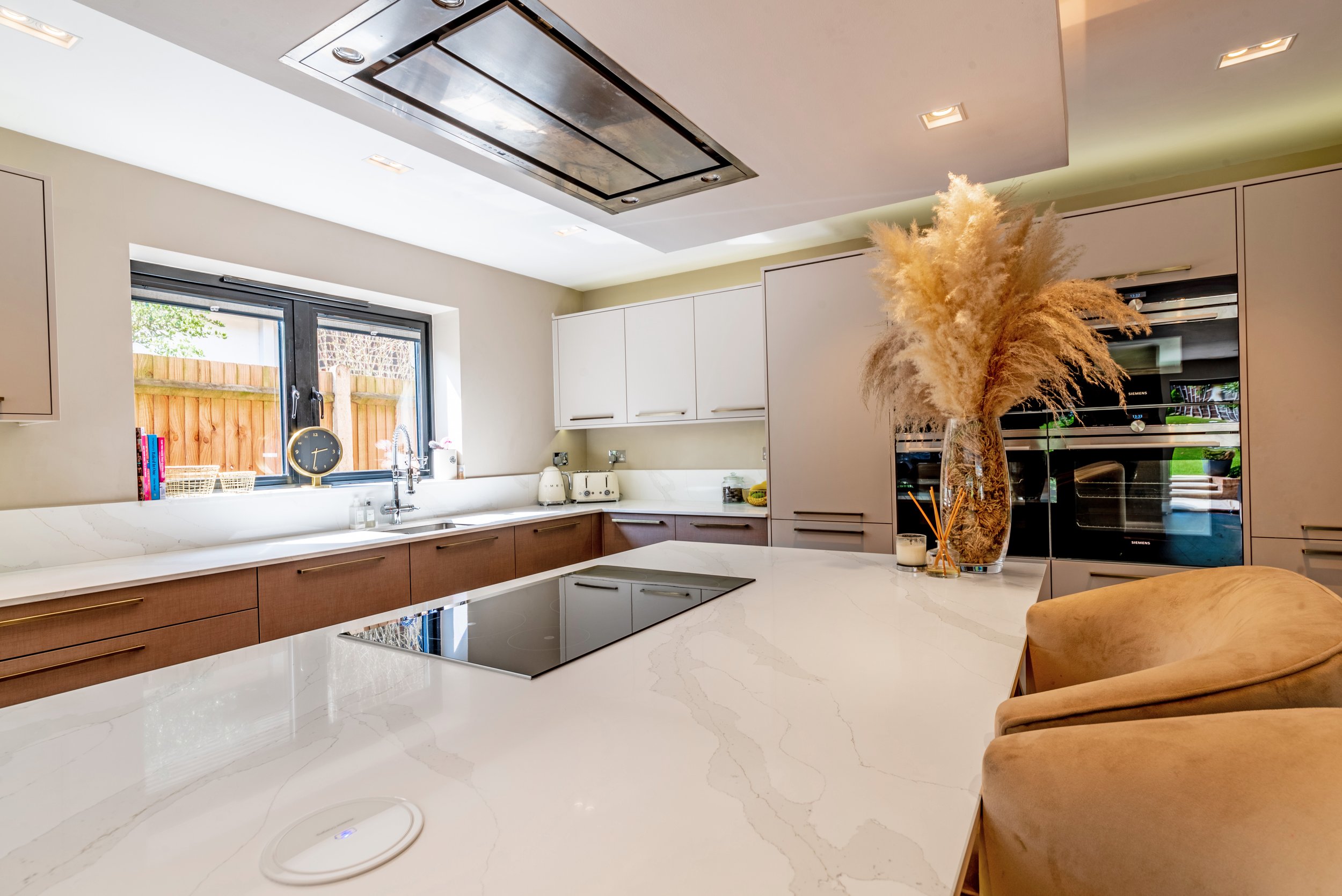 Modern kitchen with white and wood cabinetry, marble countertops, large window, and decorative vase with pampas grass.