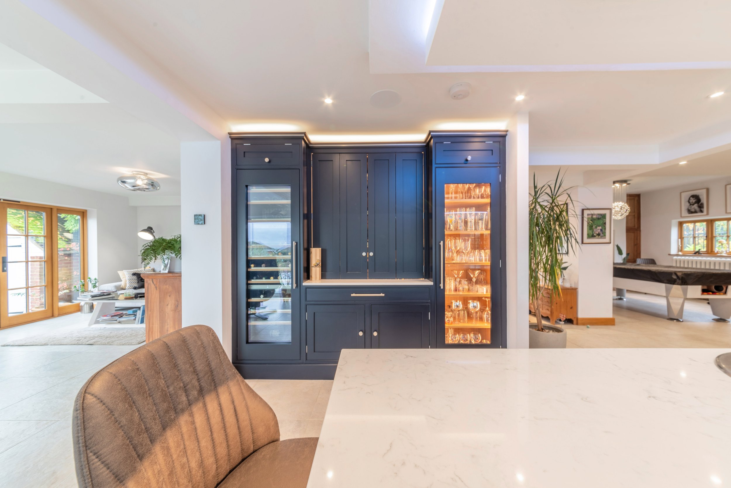 Modern home interior with bar area featuring dark cabinetry and glassware, open living space with large windows, and a pool table in the background.