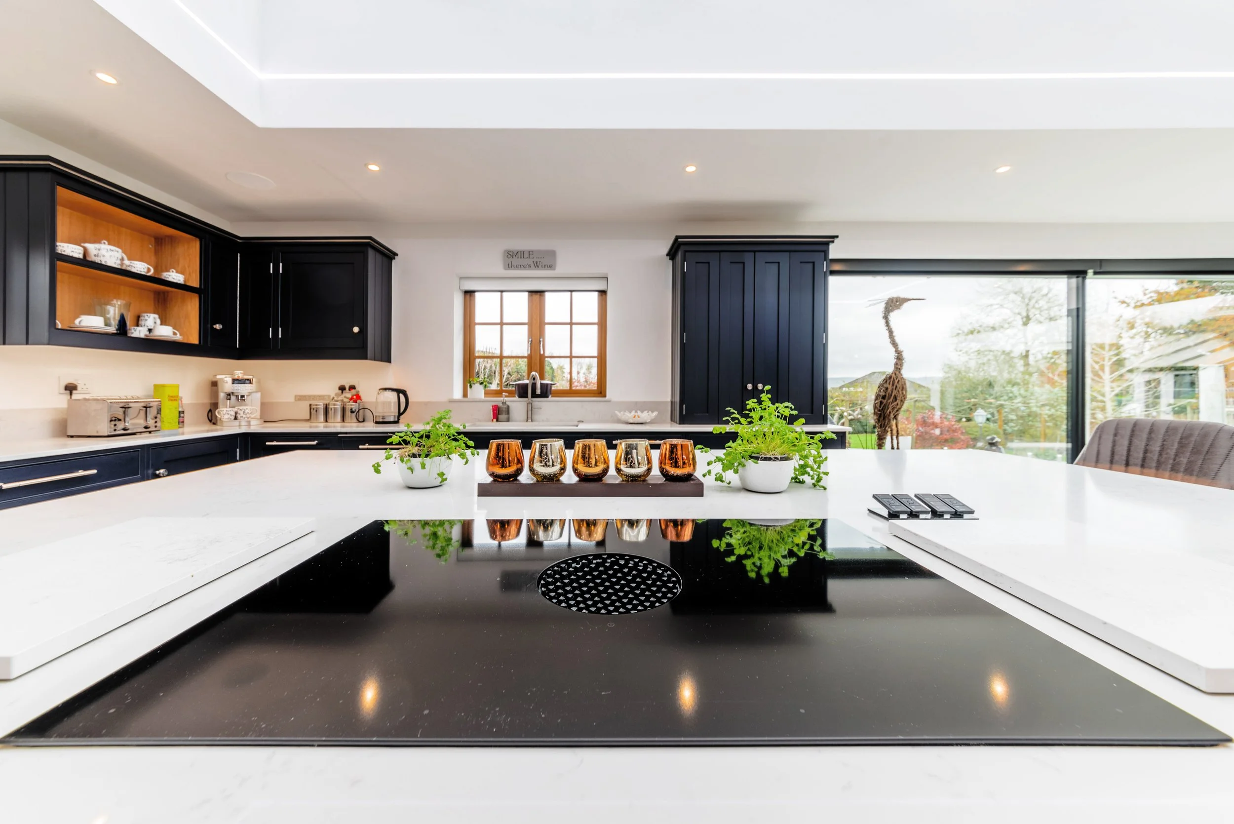 Modern kitchen with black and white cabinets, a large island, copper and gray glass cups, plants, and a giraffe outside through large windows.