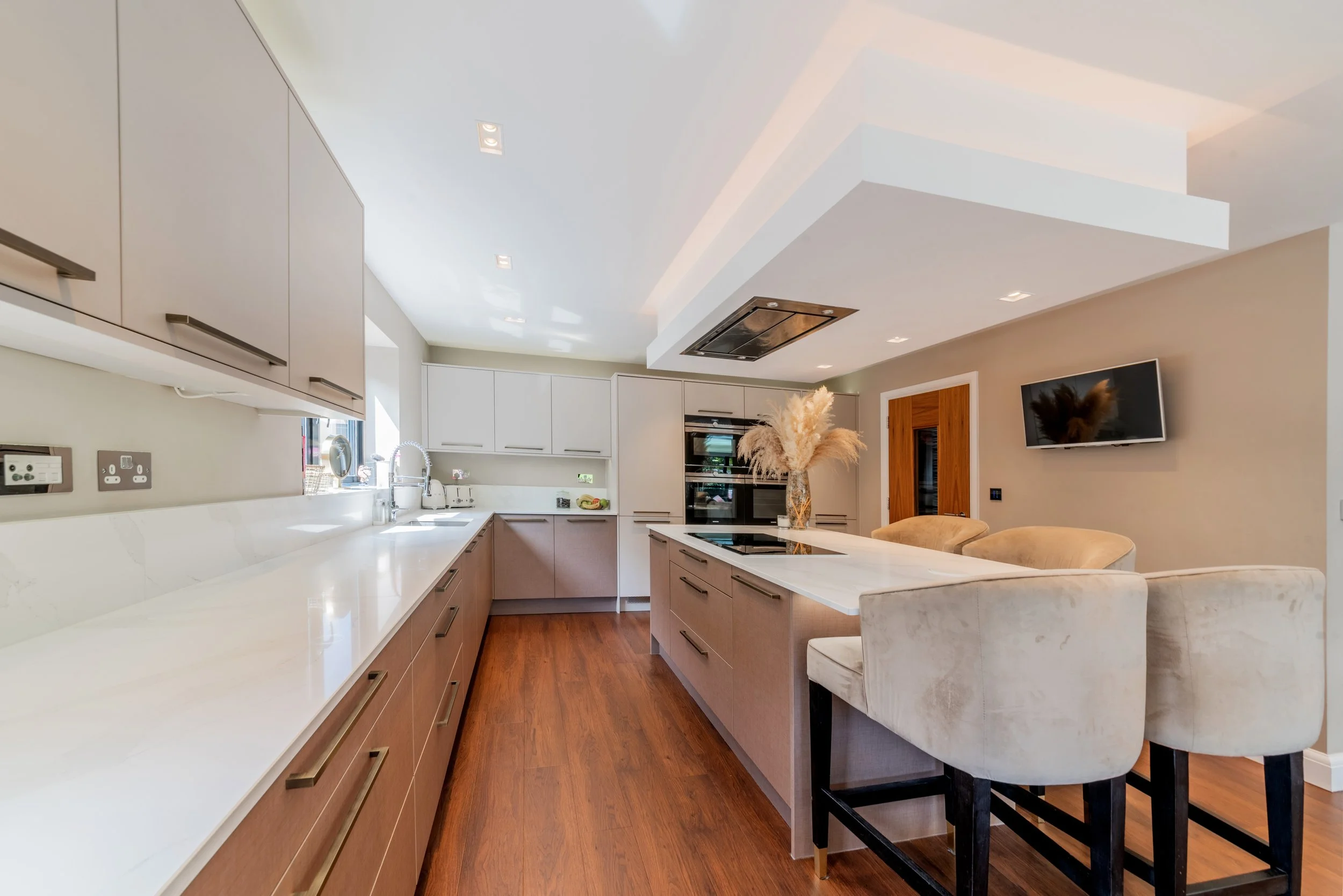 Modern kitchen with beige and white cabinets, marble countertops, and a wooden floor. Kitchen island with four beige velvet chairs, a vase with pampas grass, wall-mounted TV, and a wooden door.