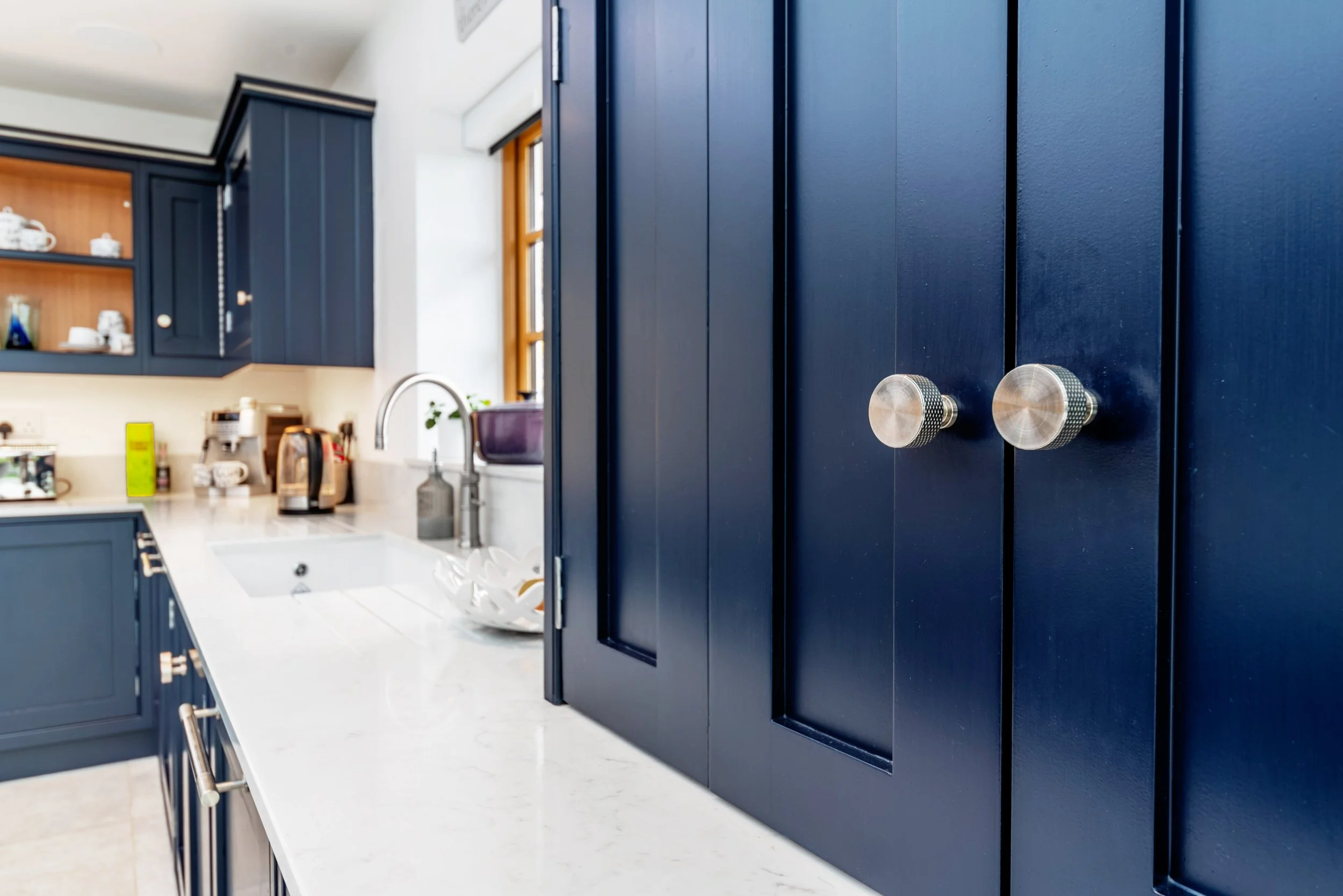 Close-up of blue kitchen cabinets with metallic knobs, part of a modern kitchen with a white marble countertop, sink, and window.
