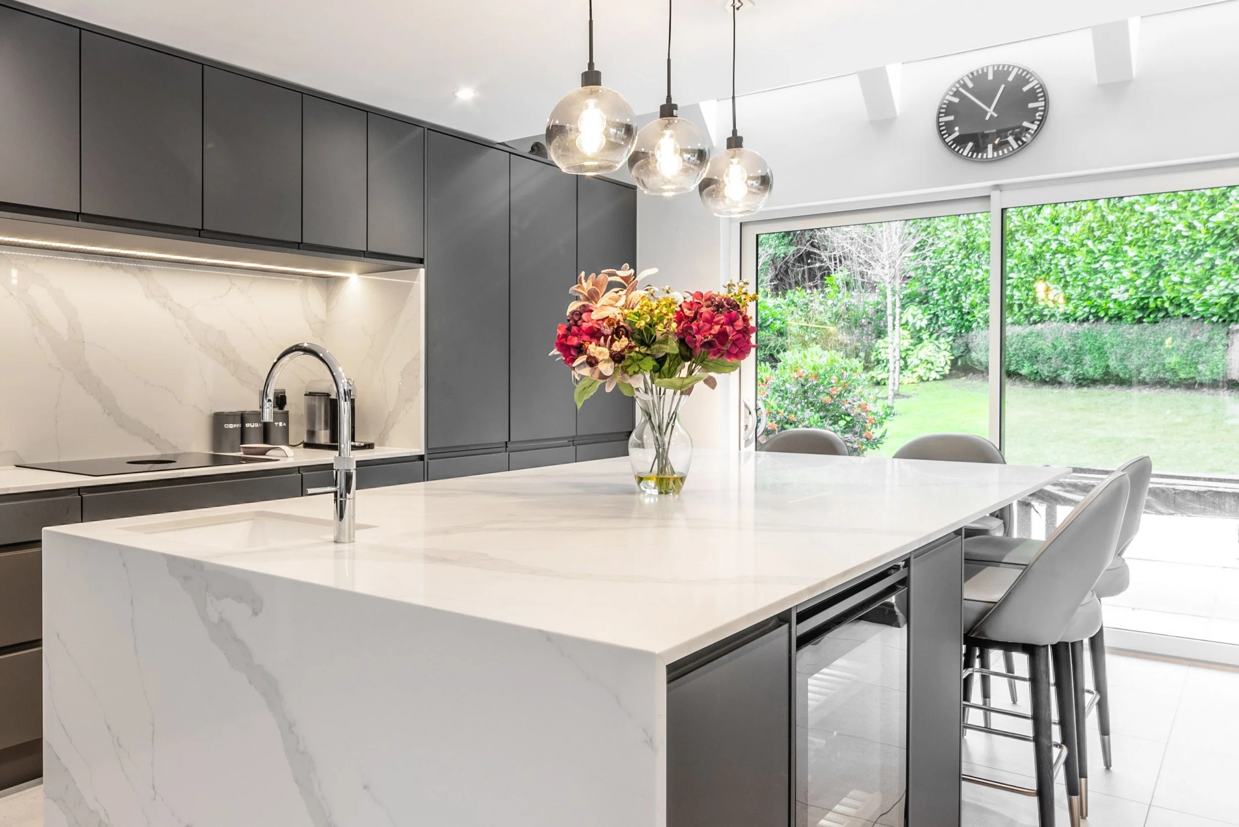 Modern kitchen with white marble island, dark gray cabinets, flower arrangement on the counter, large window showing a garden, three pendant lights, and a clock on the wall.
