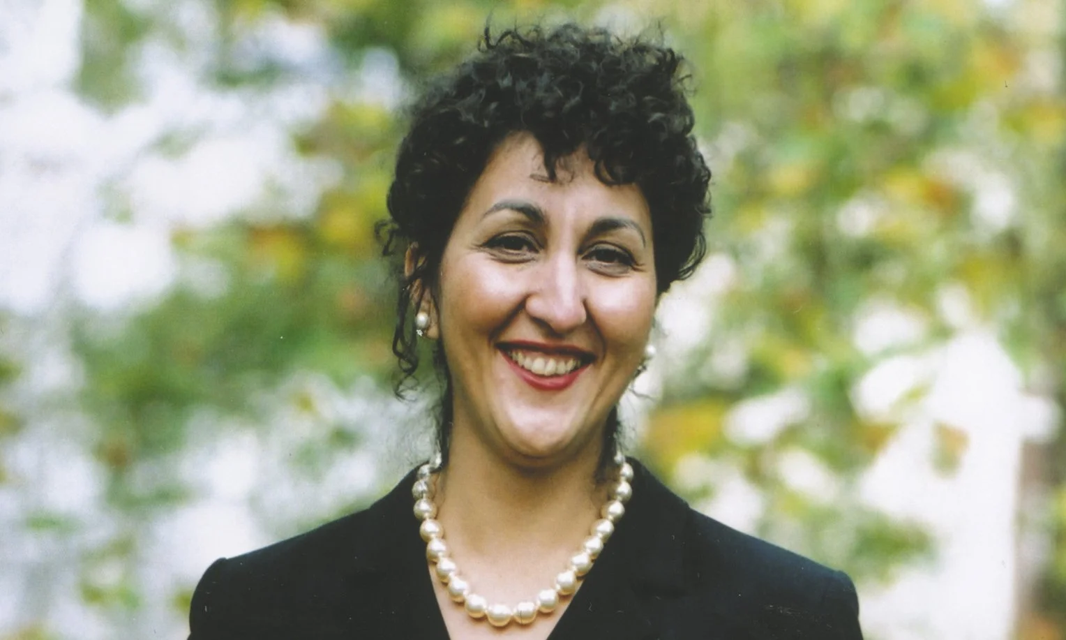 A woman with short curly black hair wearing pearl earrings and necklace, smiling in an outdoor setting with green foliage in the background.