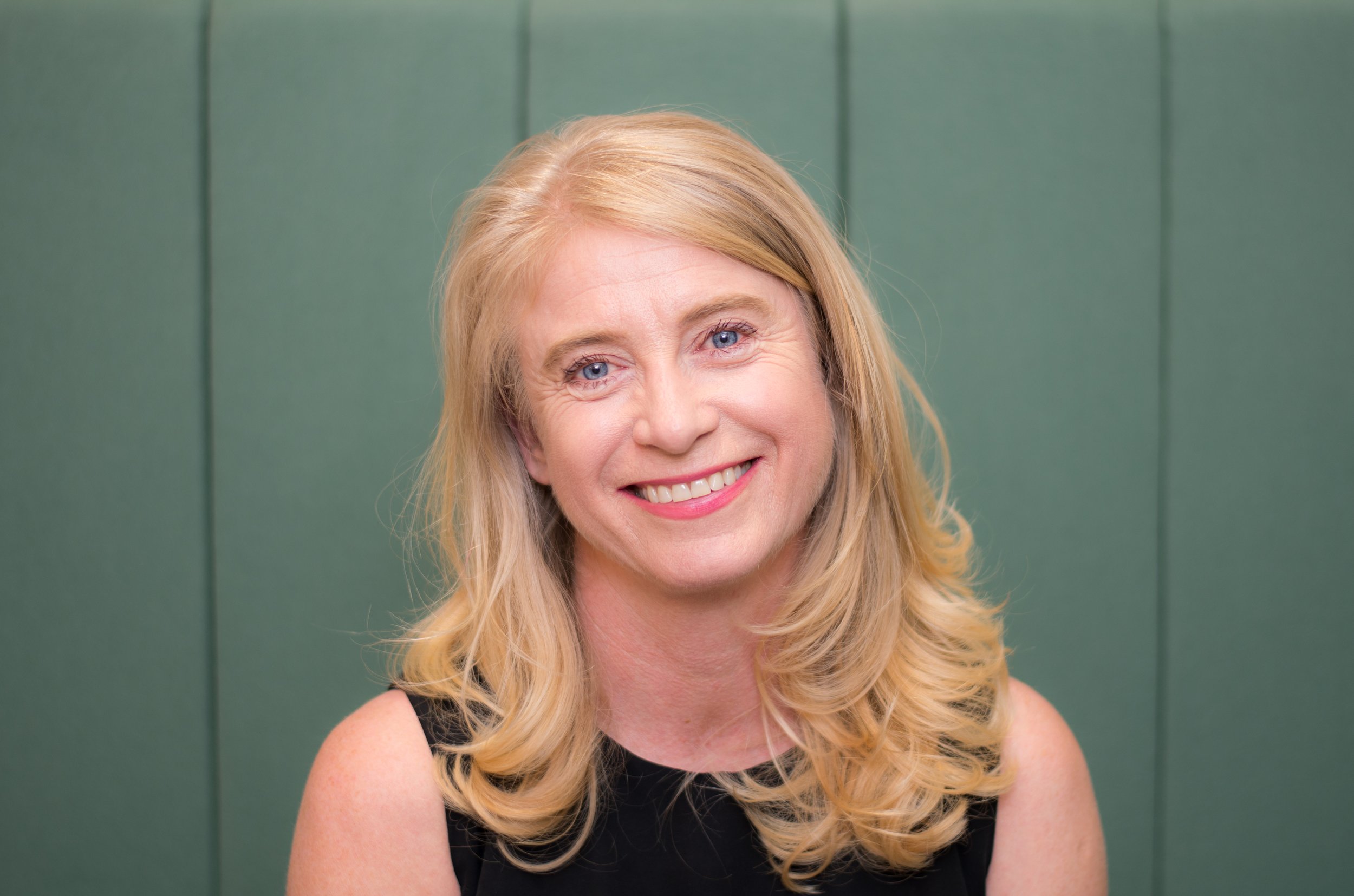 Smiling woman with blonde hair and blue eyes, wearing a black sleeveless top, standing against a green background.