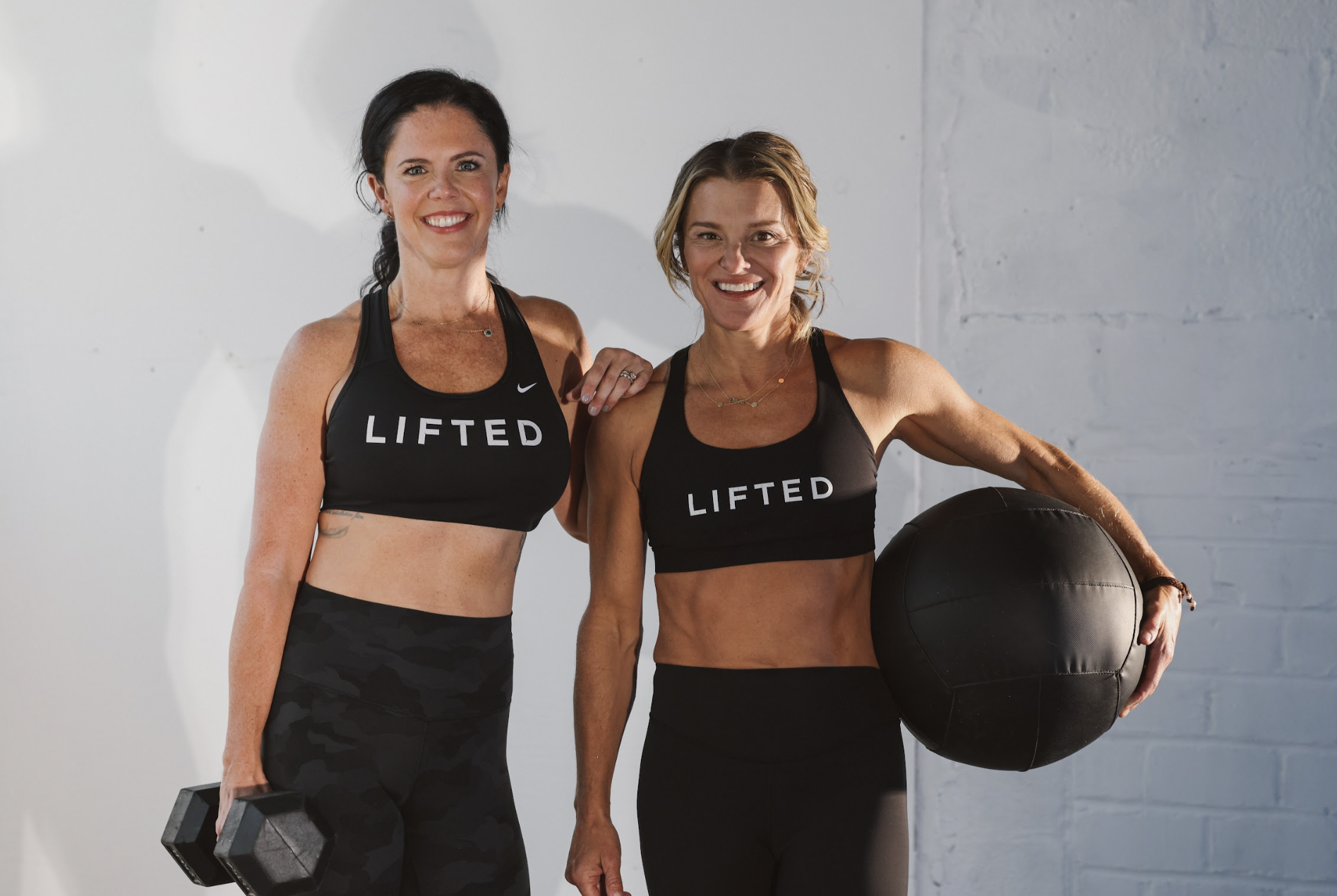 Two women in workout clothes smiling at the camera, one holding a dumbbell and the other holding a medicine ball, in a gym setting.
