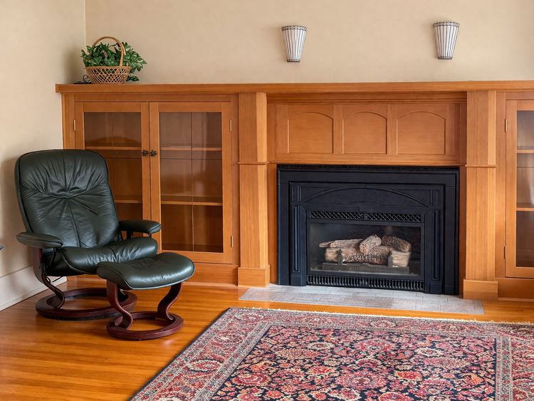 A living room corner with a black leather armchair, a wooden cabinet with glass doors, a fireplace, and a decorative rug.